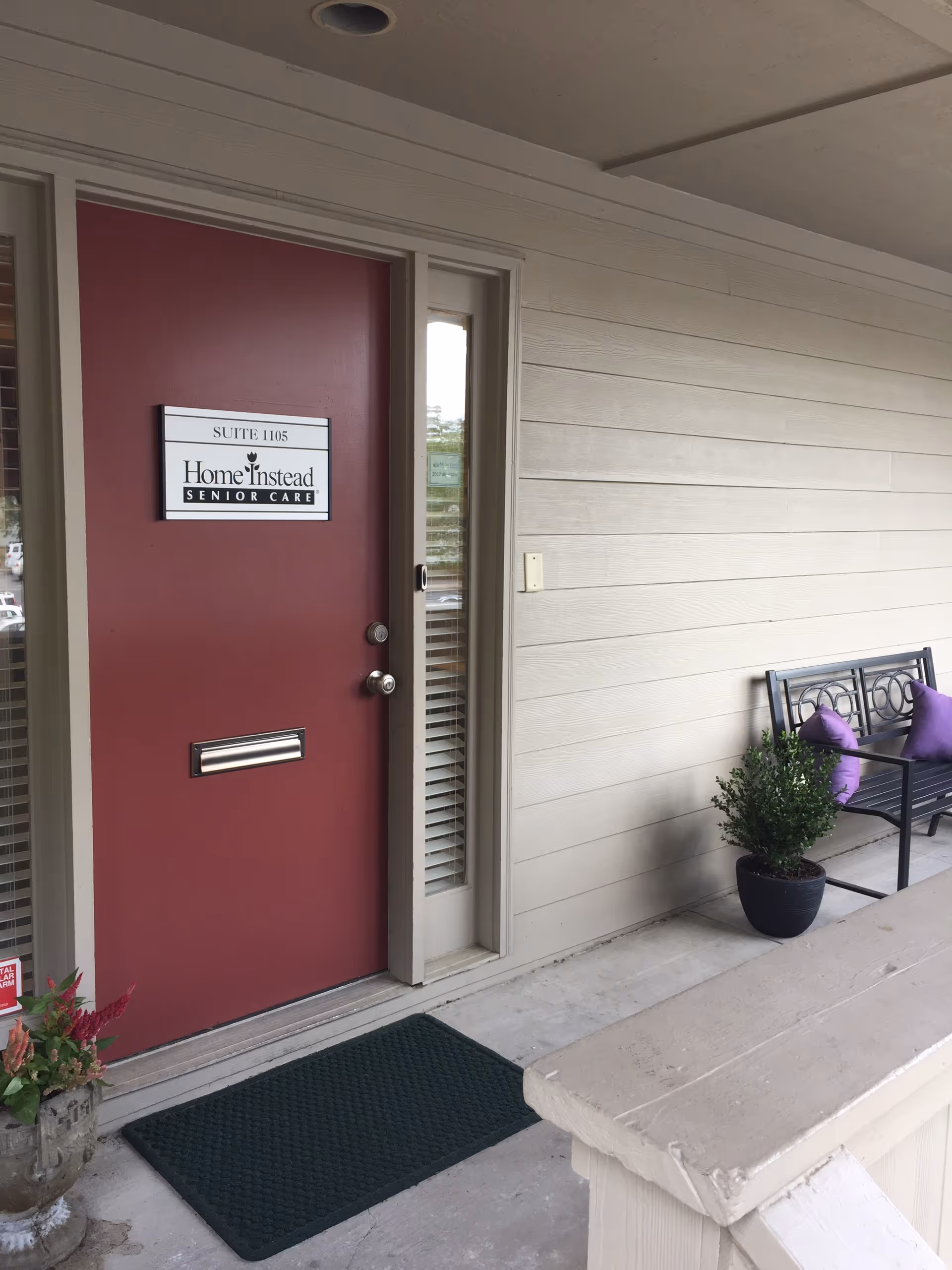 Red door with Home Instead Senior Care sign, beige siding, and a bench outside.