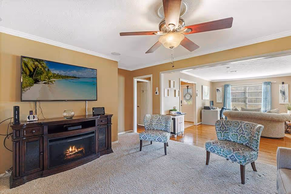 Living room area with fireplace, mounted TV, ceiling fan, and two patterned chairs.