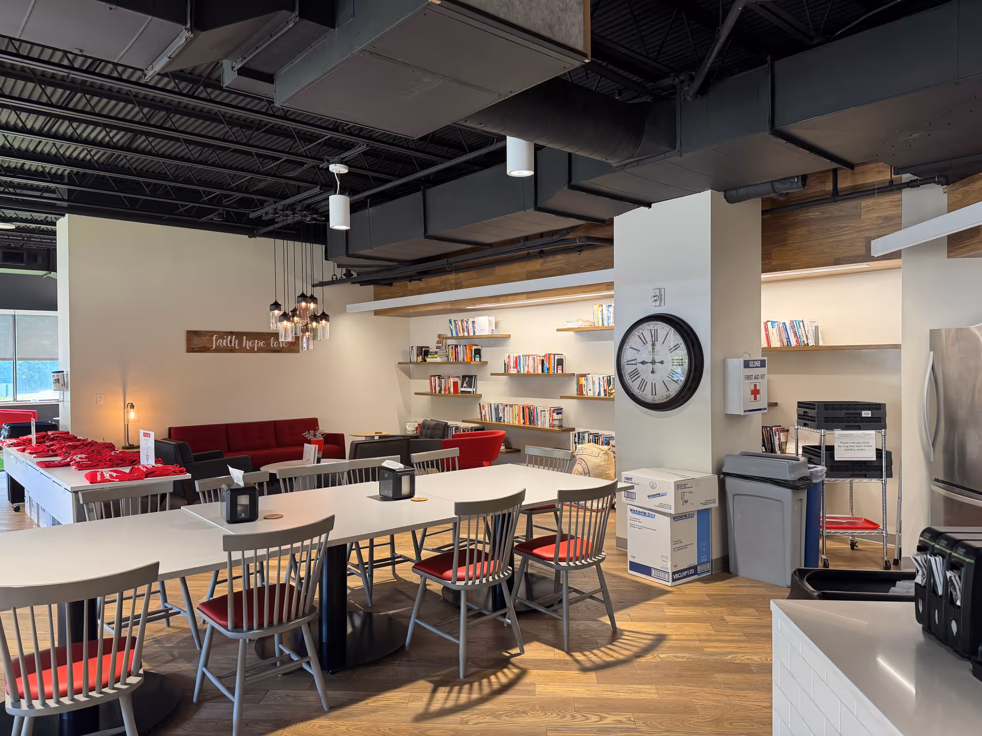 Interior community room with long tables, metal chairs, a bookshelf wall, clock, and kitchen area.