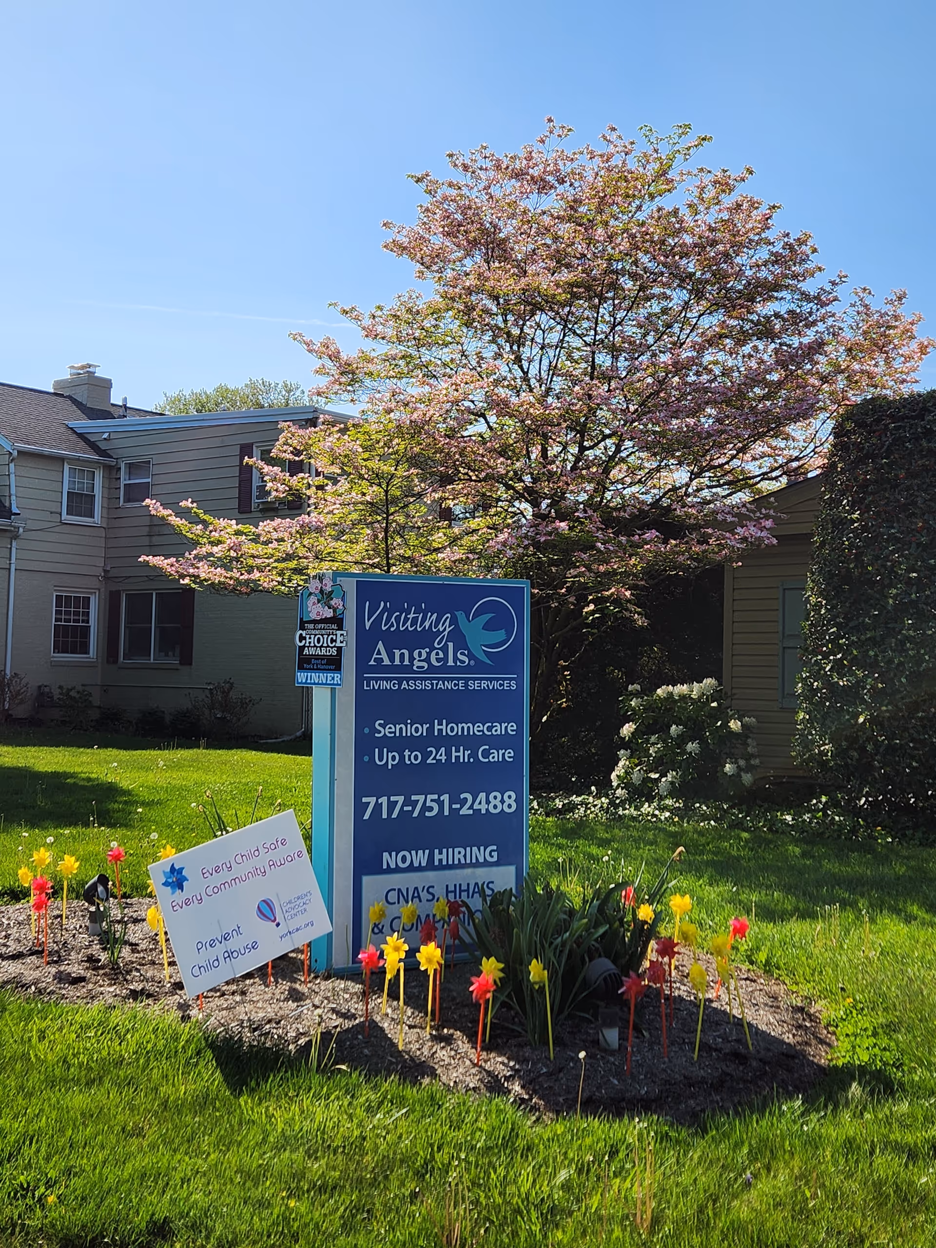 Blue Visiting Angels sign in a sunny front yard with blooming tree and flowers.