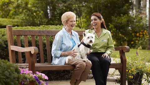 An elderly woman and caregiver sit on a wooden outdoor bench, smiling with a small dog.