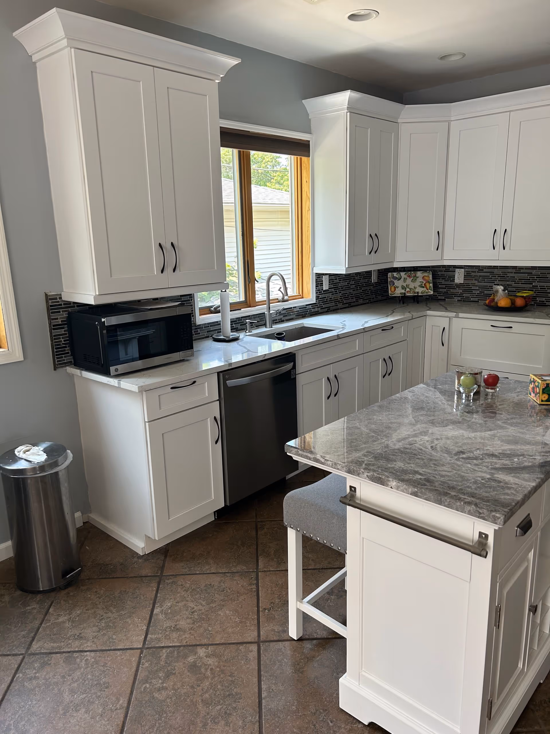 White kitchen with marble island, stainless appliances, tiled floor, and sunny window.