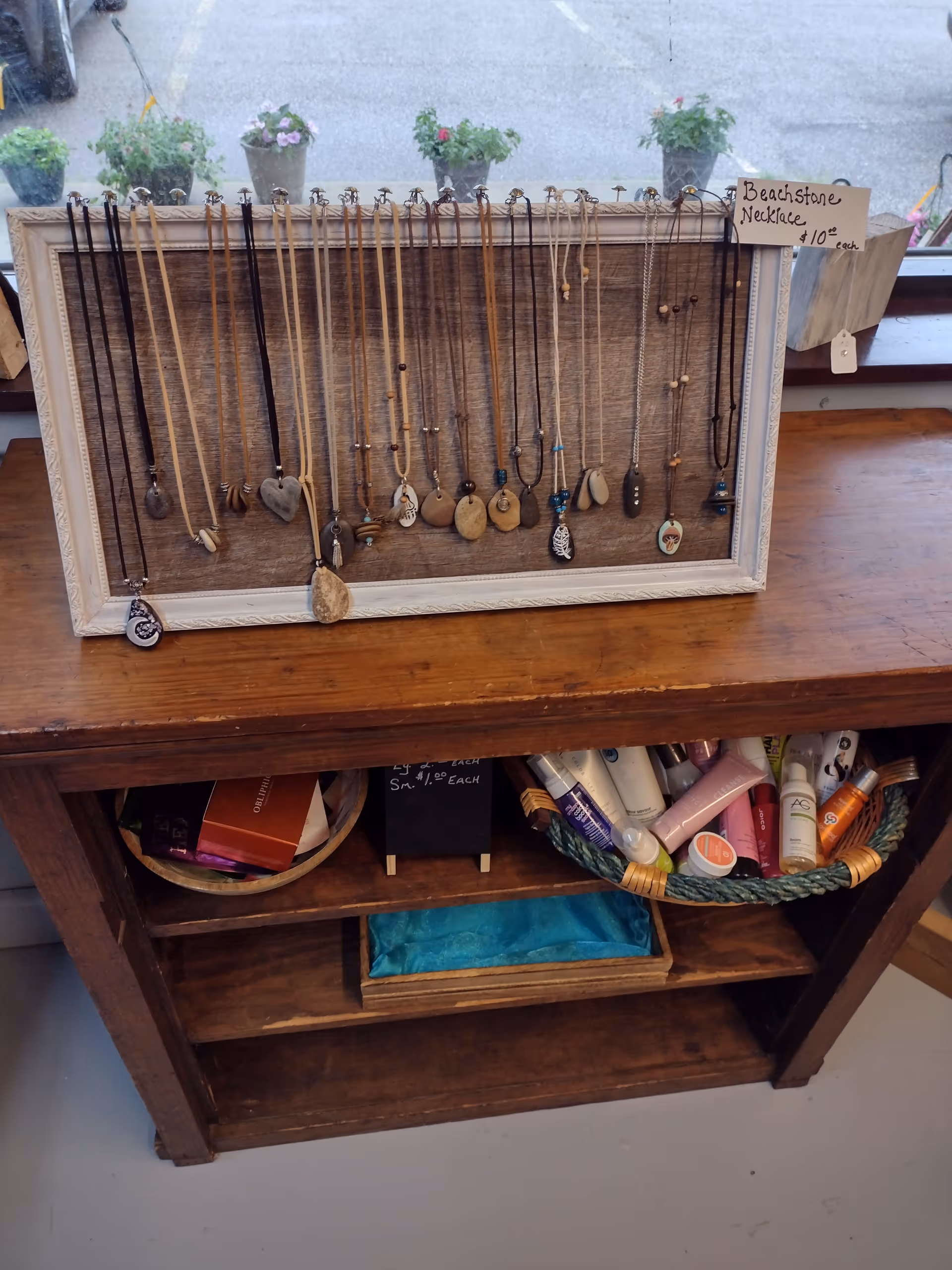 A wooden table display showcasing assorted necklaces with a small sign and plants in background.