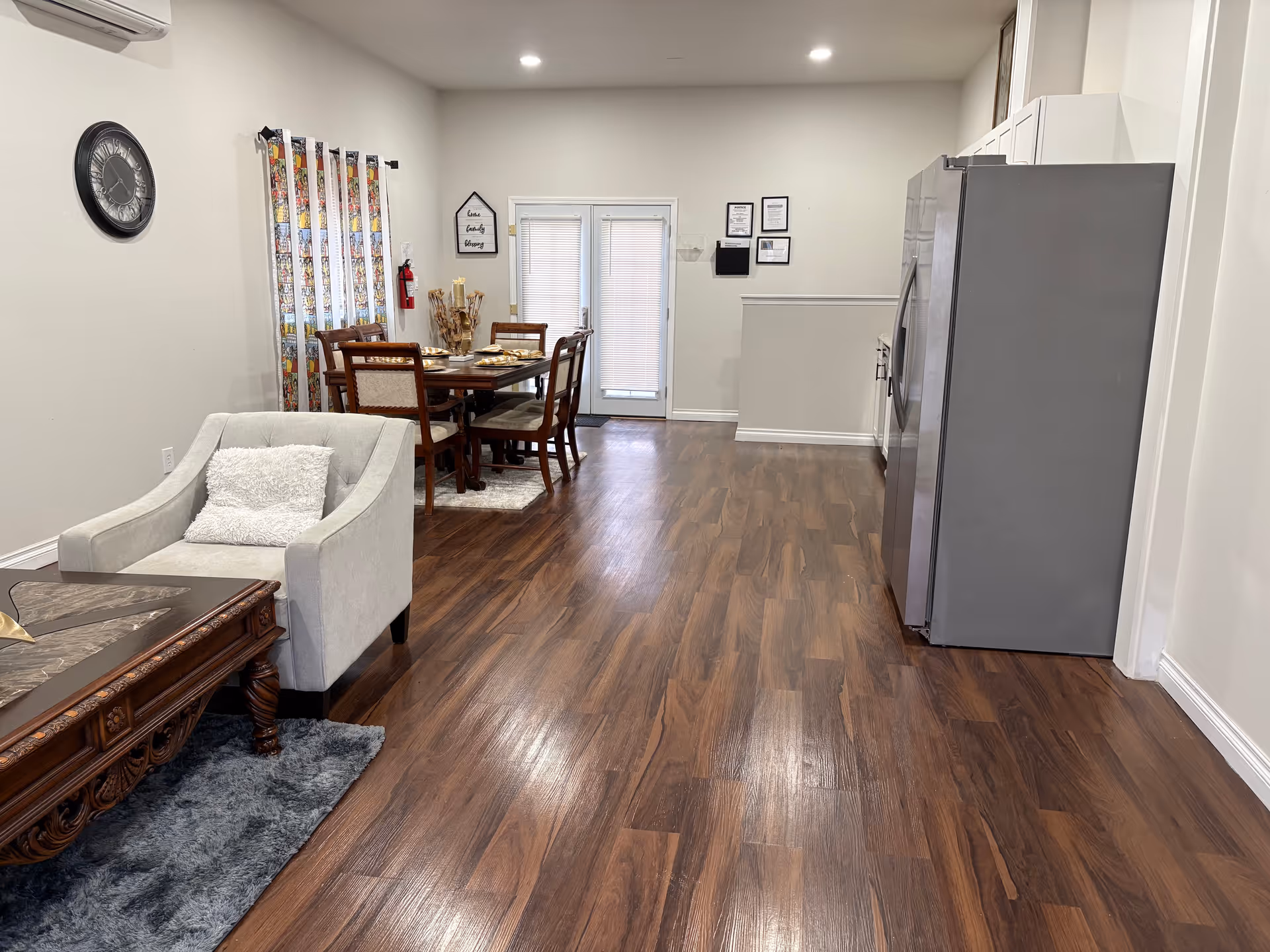 Open-plan senior living common area with a sofa, dining table, and large refrigerator.