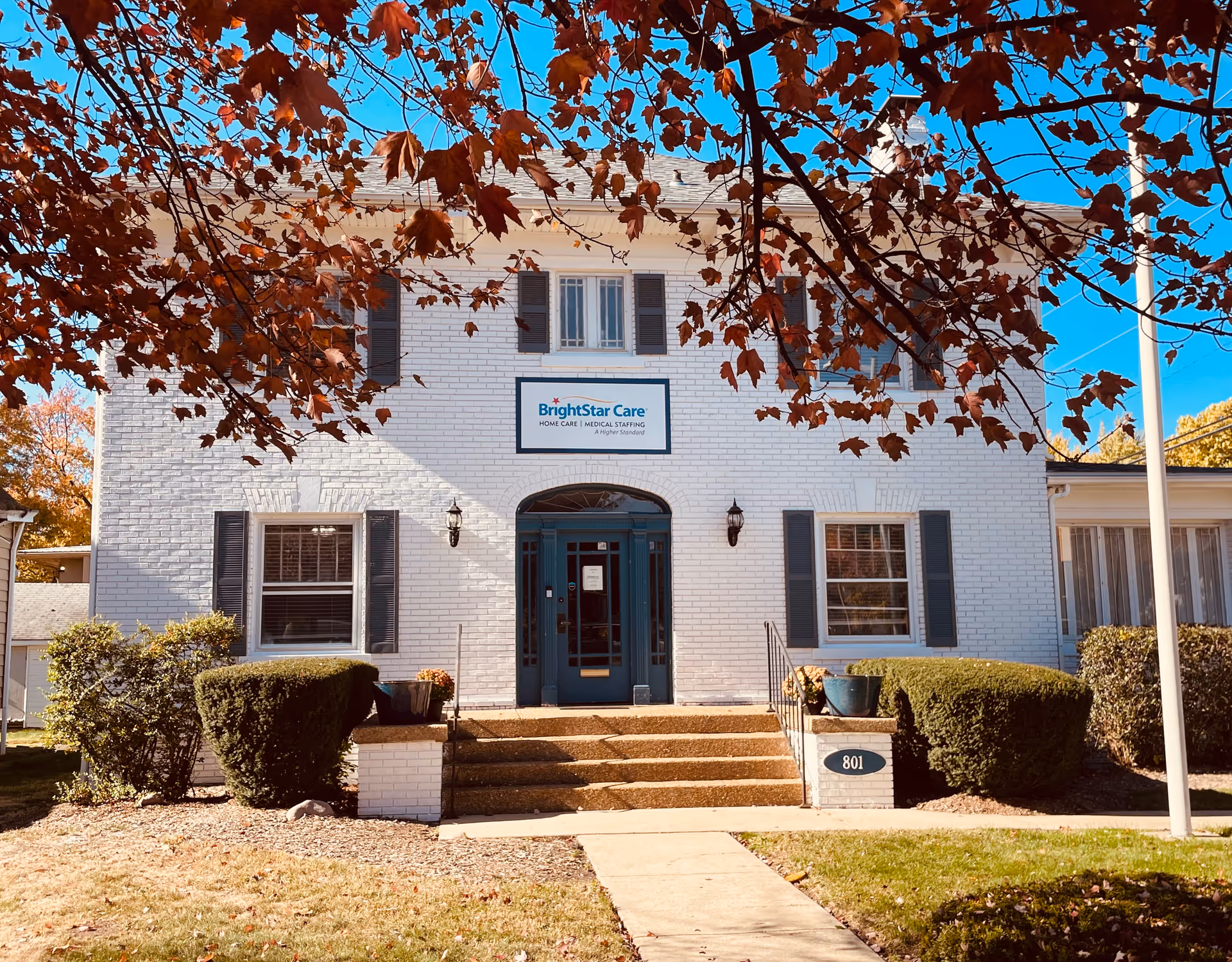 White brick BrightStar Care building with blue entrance, steps, shrubs, and autumn leaves overhead.