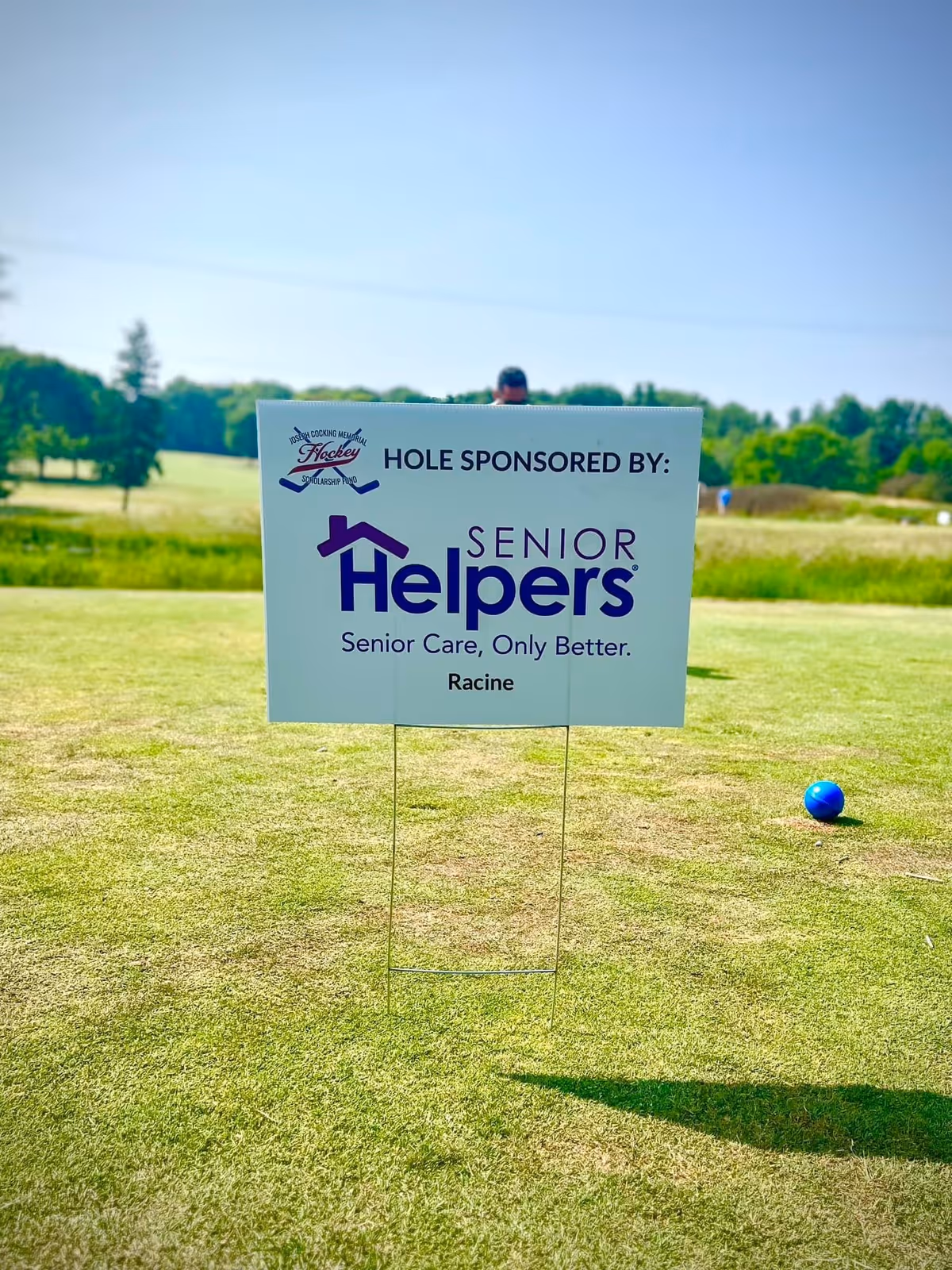 Outdoor golf course with a Senior Helpers sponsor sign and a distant golfer in background.