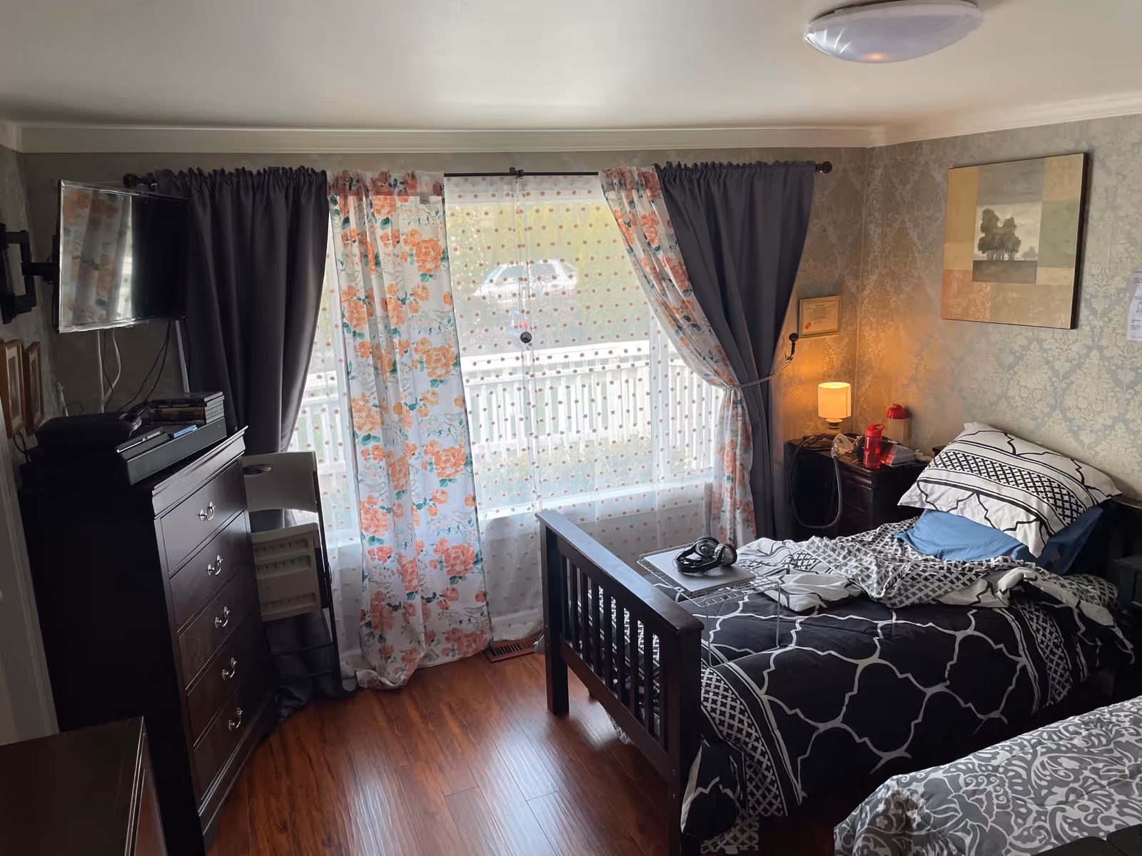A furnished bedroom in a senior living facility with two beds, dresser, TV, and floral curtains.