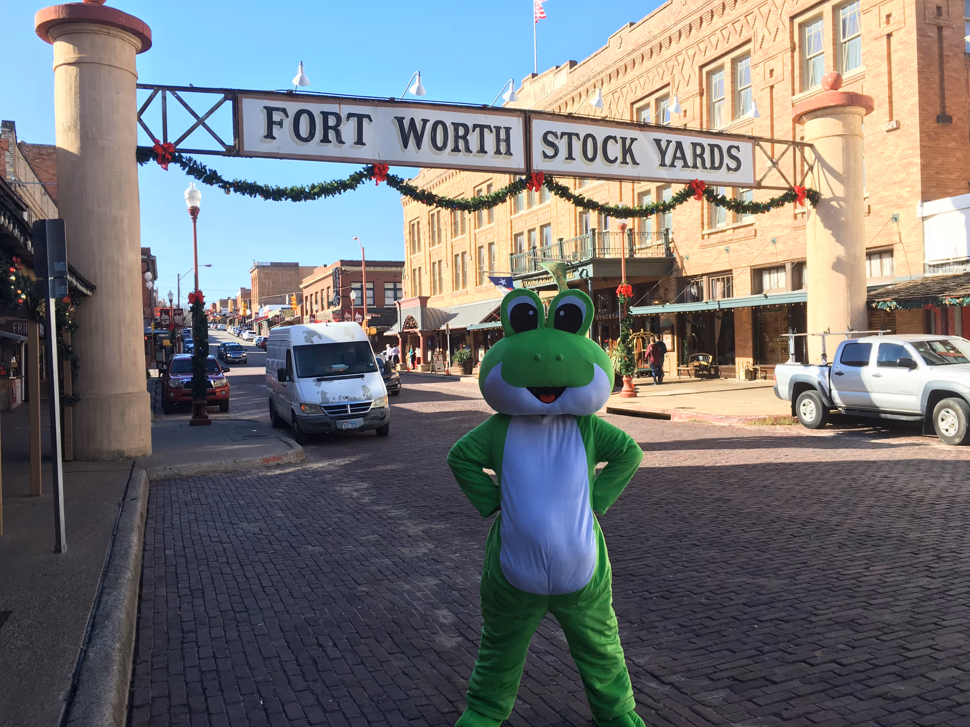 Green frog mascot stands on a brick street under the Fort Worth Stock Yards sign.