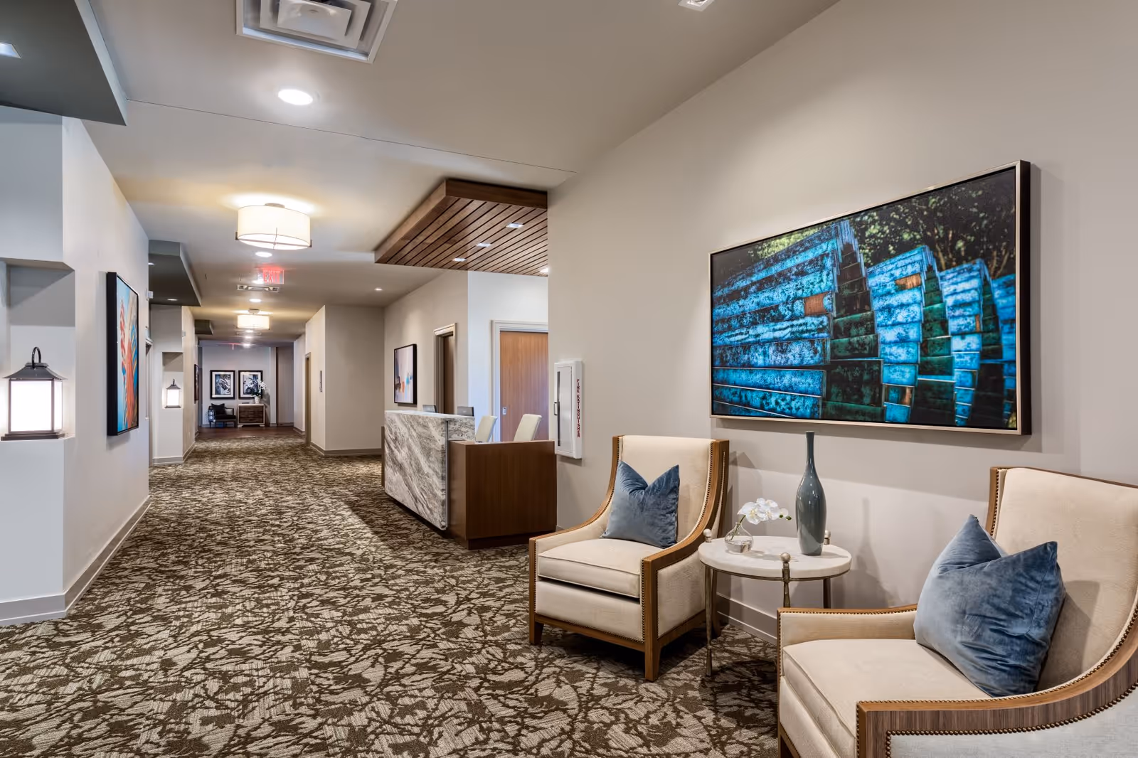 Interior lobby with two beige armchairs, a round table, artwork, and a wall-mounted TV.