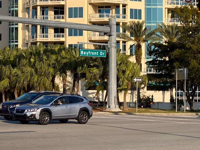 Cars drive past a modern building with palm trees and a Bayfront Dr street sign.