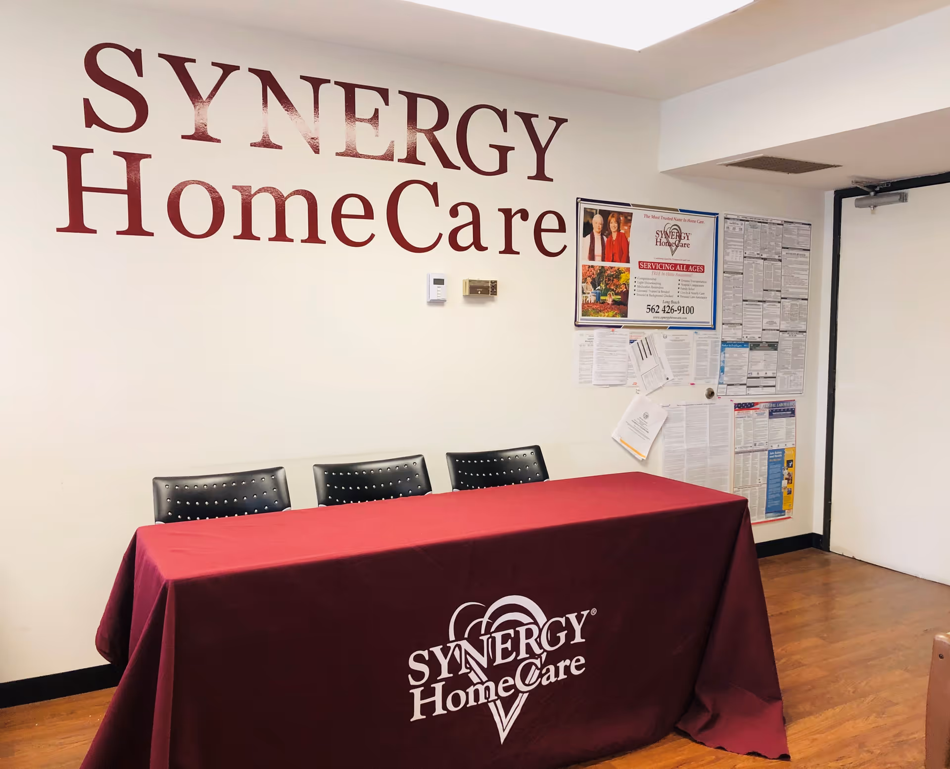 Interior reception area with large SYNERGY HomeCare sign on wall, maroon table, three chairs.