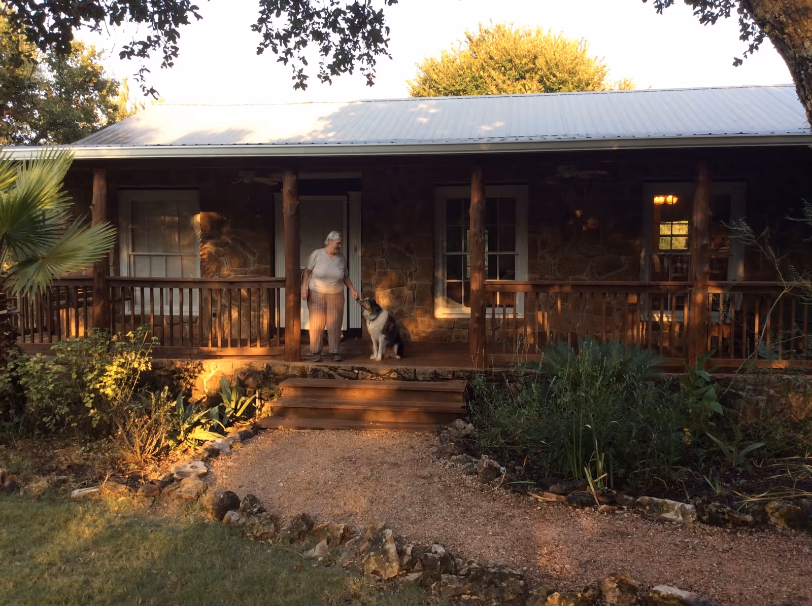 An elderly woman stands on a stone house porch with a leashed dog beside her.