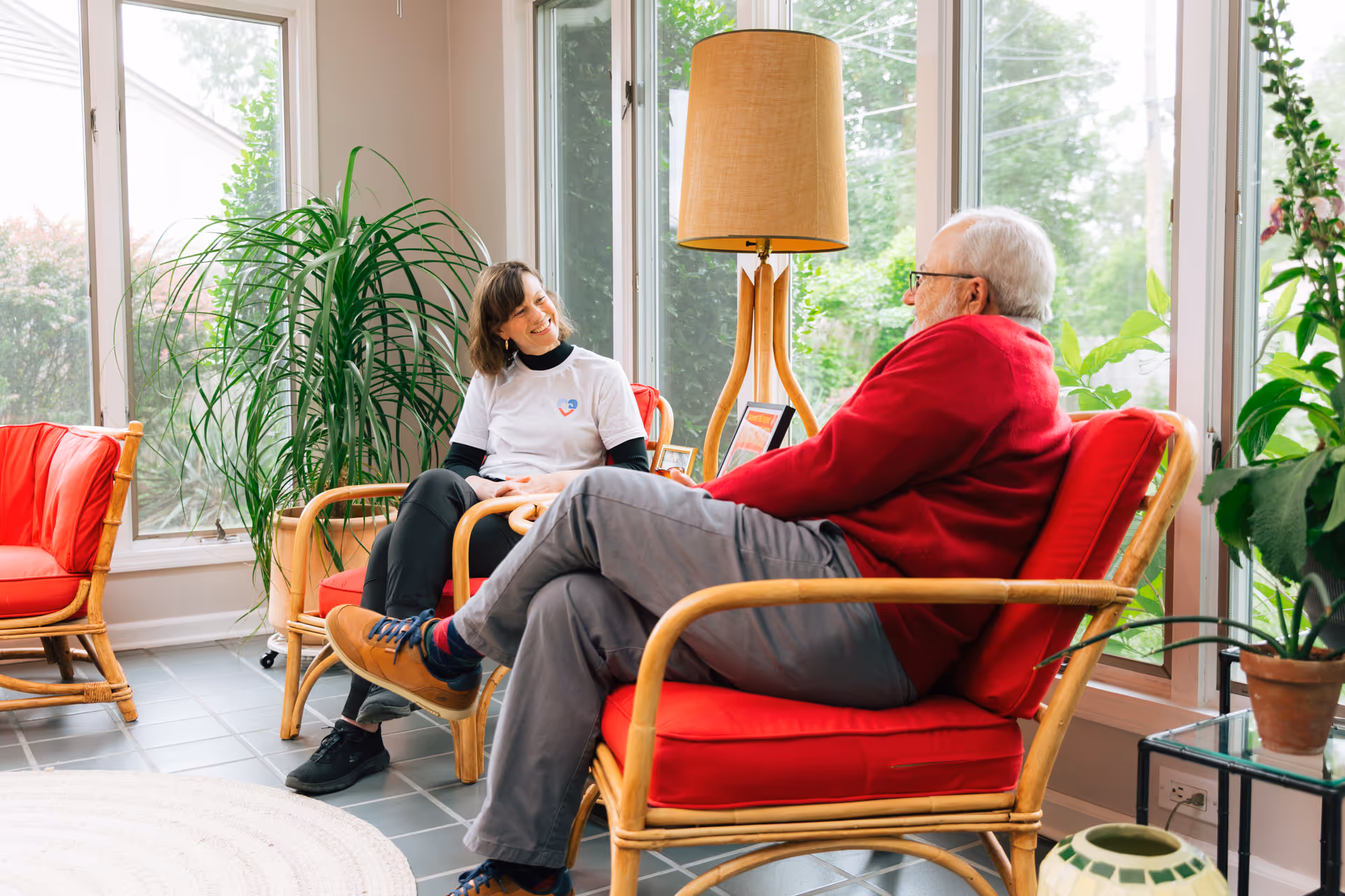 Two seniors sit and chat in a bright sunlit living room with red cushioned chairs.