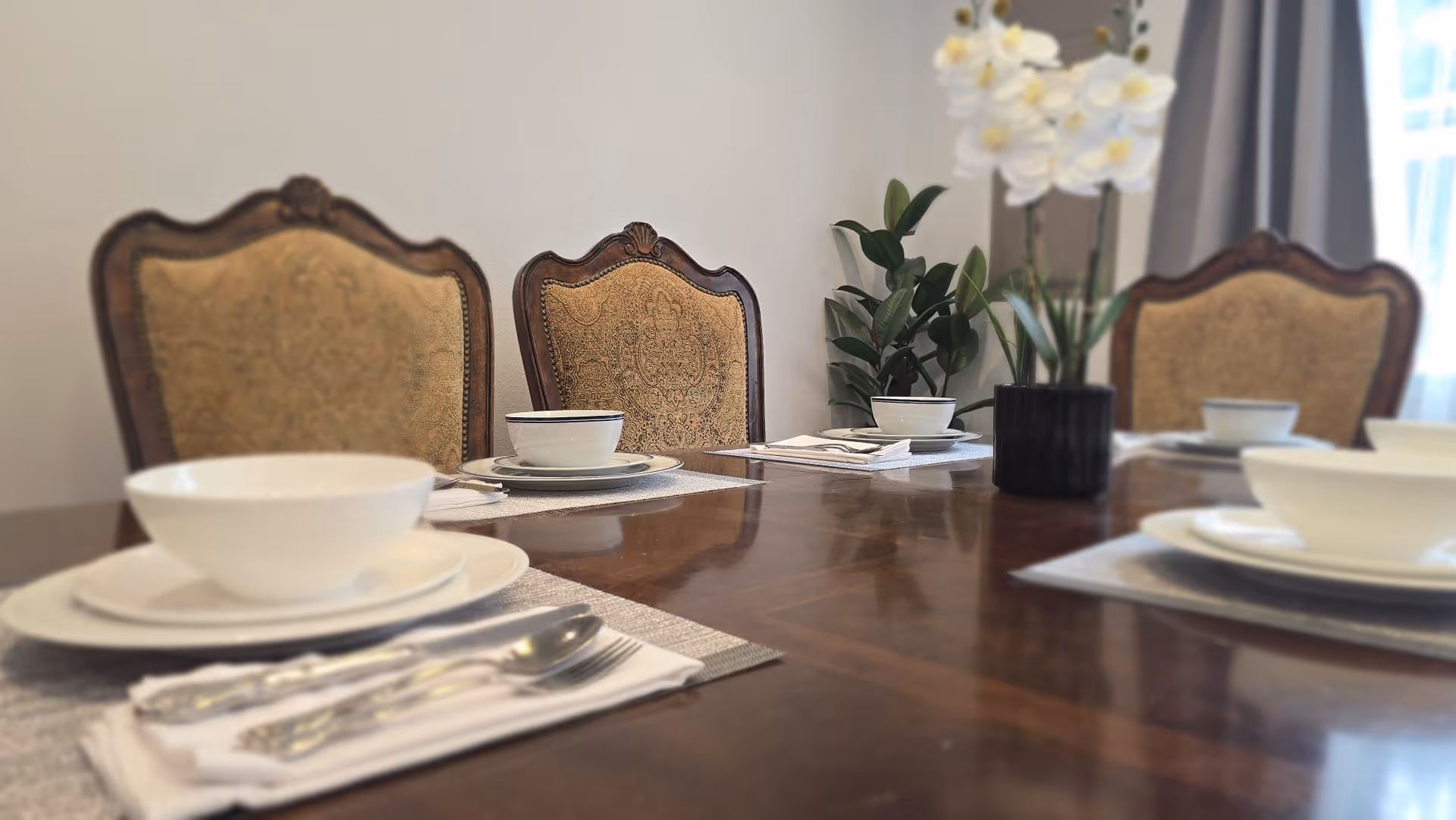 A dining table is fully set with bowls, plates, utensils, and an orchid centerpiece.