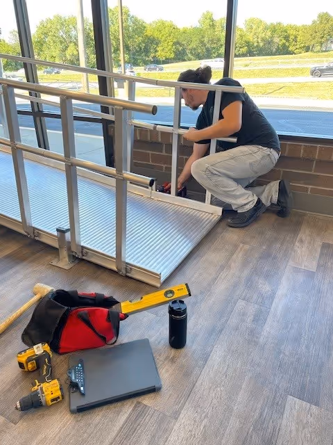 Maintenance worker kneels beside a wheelchair lift by large interior windows.