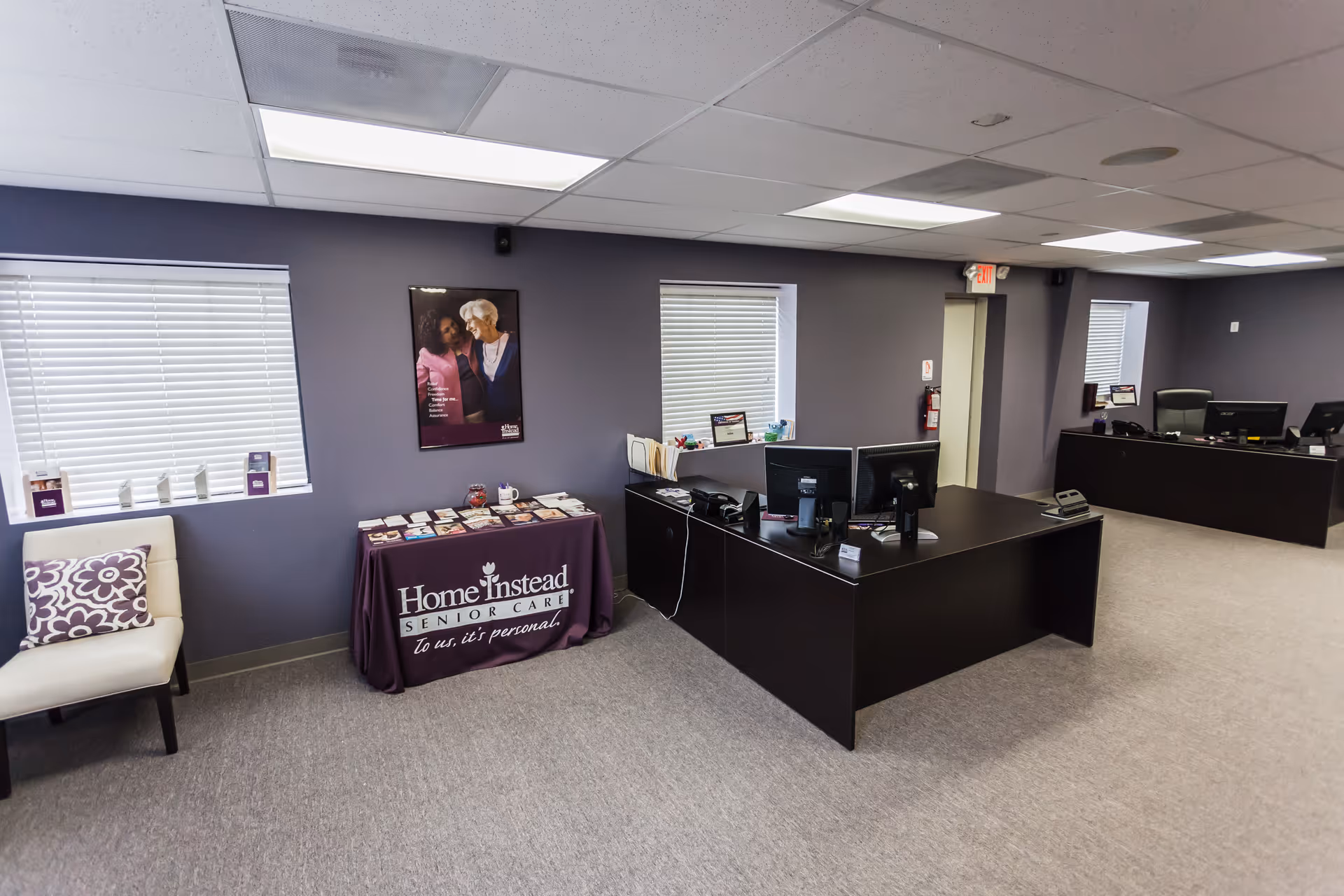 Office reception area with Home Instead branding, computer desks, and seating along purple walls.