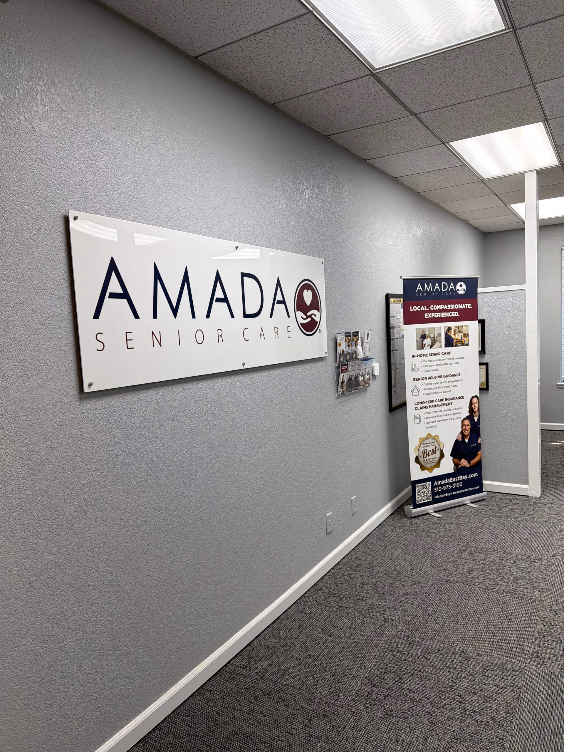 Interior hallway with AMADA Senior Care sign and promotional display along a gray wall.