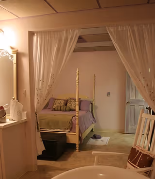 Canopy bed with sheer curtains in a pastel bedroom, vanity mirror on left, purple bedding.