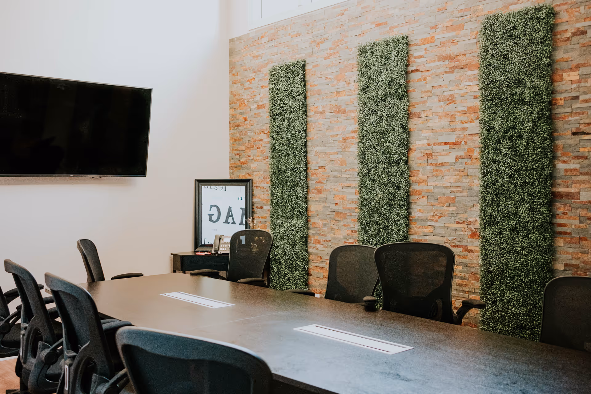 A long conference table with black chairs in a meeting room with brick wall and greenery.