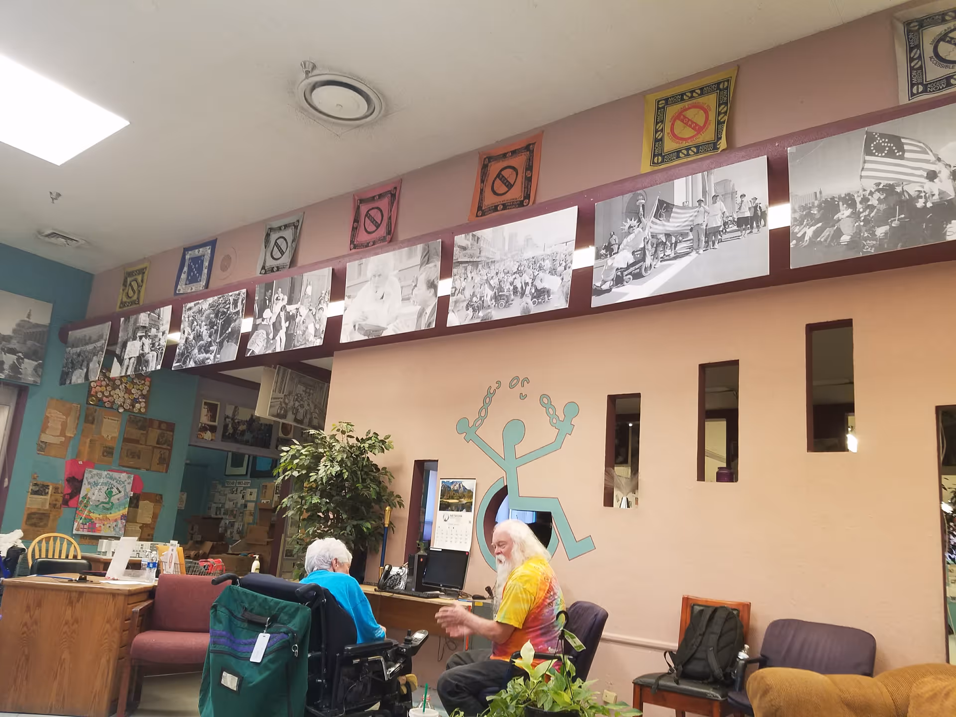Two elderly residents sit at a desk in a bright communal interior with wall art.
