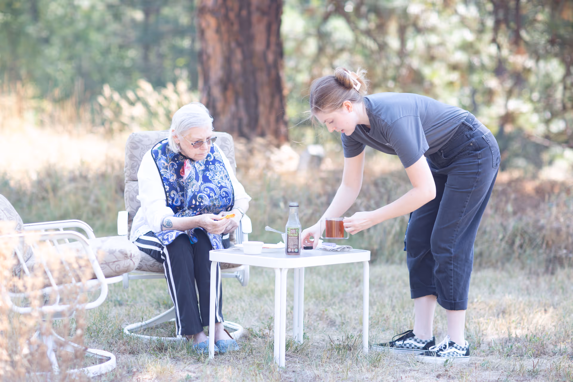 Caregiver serves a beverage to an elderly woman seated outdoors at a small table.