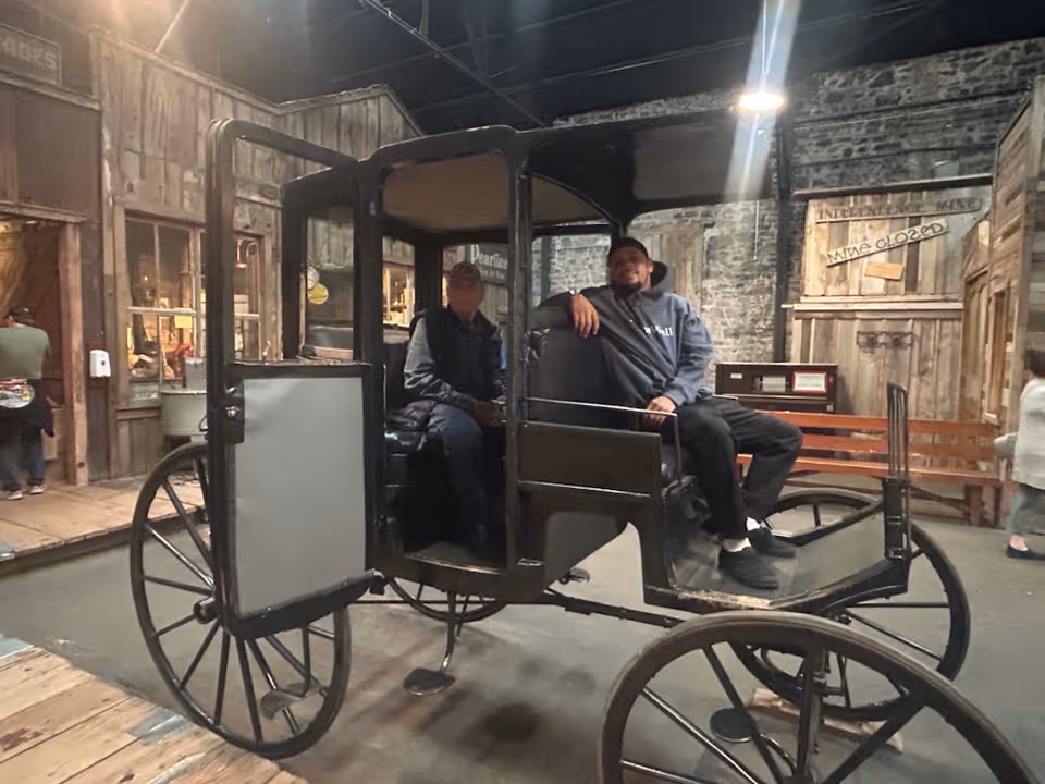 Two people sit in a vintage horse-drawn carriage inside a rustic museum exhibit