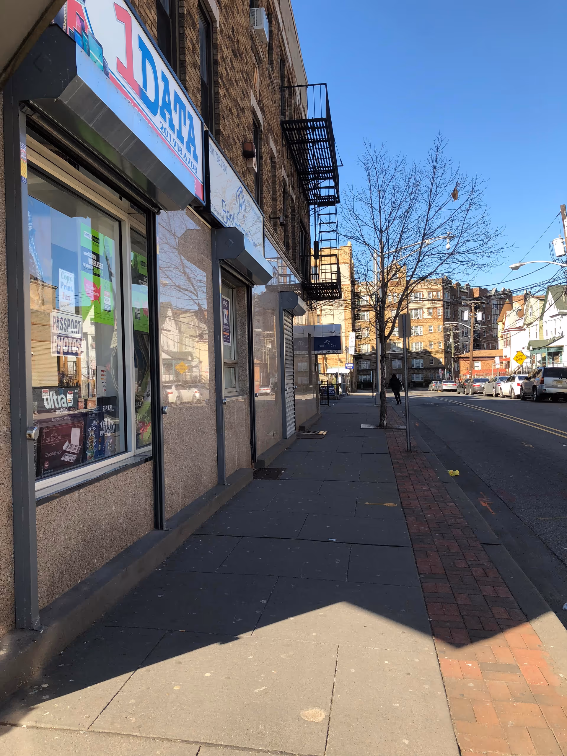 Urban street view with storefronts, brick buildings, and bare trees alongside the sidewalk.