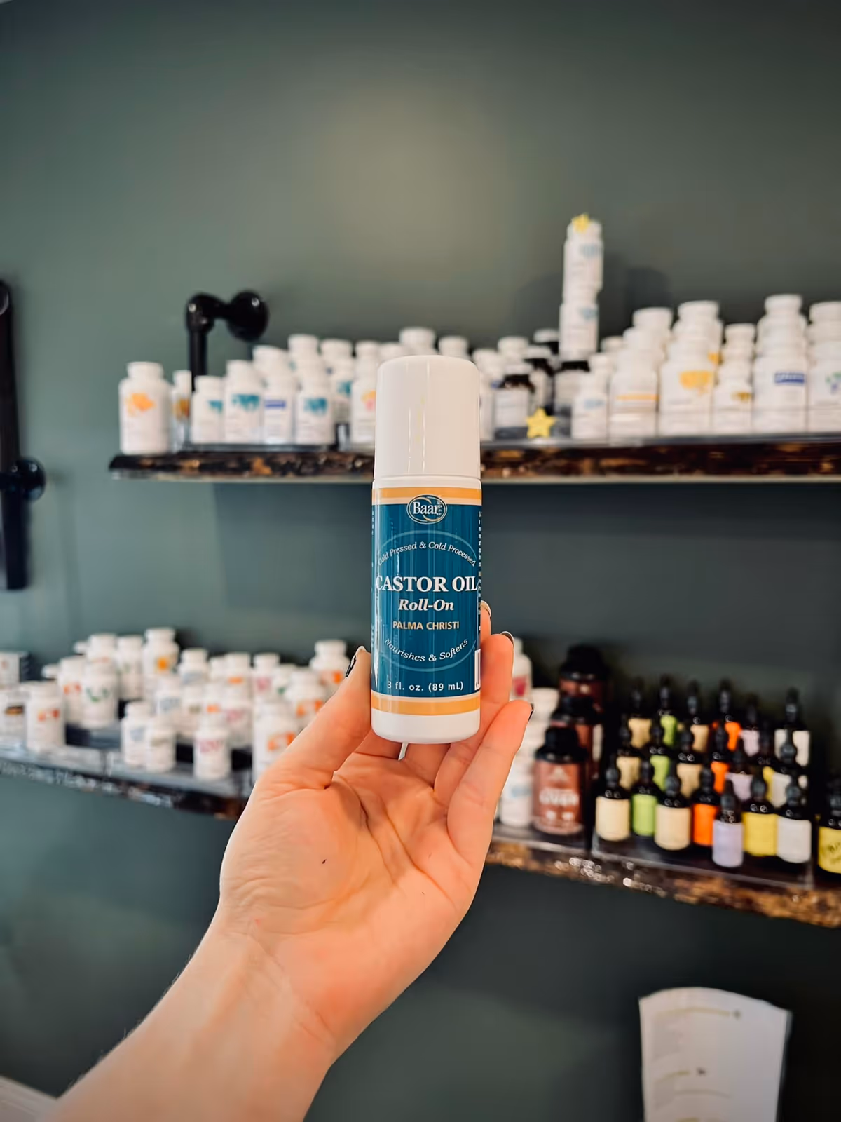 Hand holds castor oil bottle with shelves of bottles behind.
