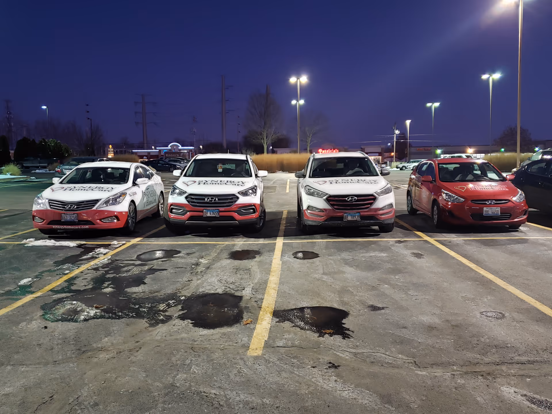 Nighttime exterior parking lot with four branded SYNERGY HomeCare cars lined up.