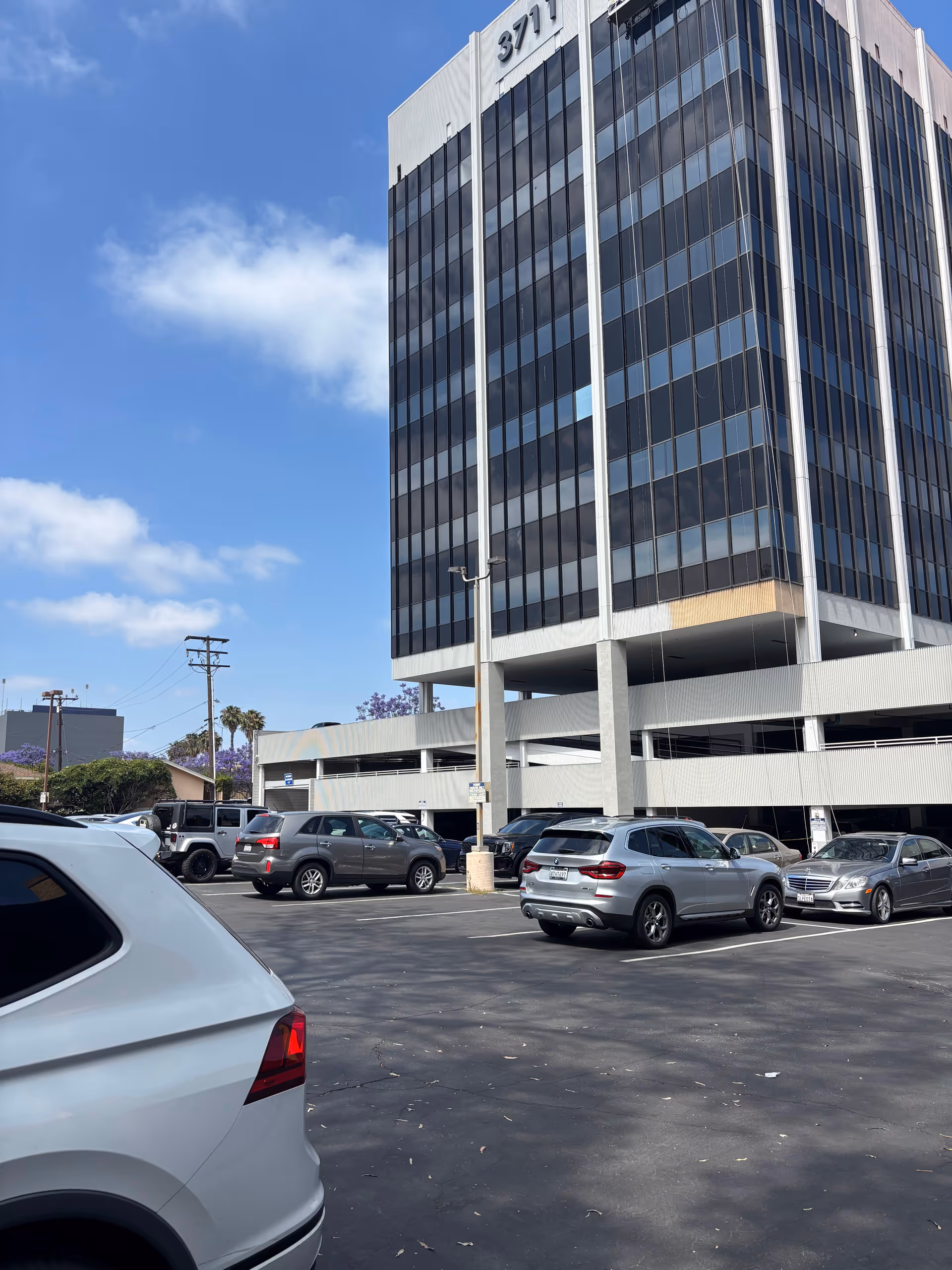 Tall glass office building with a multi-level parking structure and several parked cars.