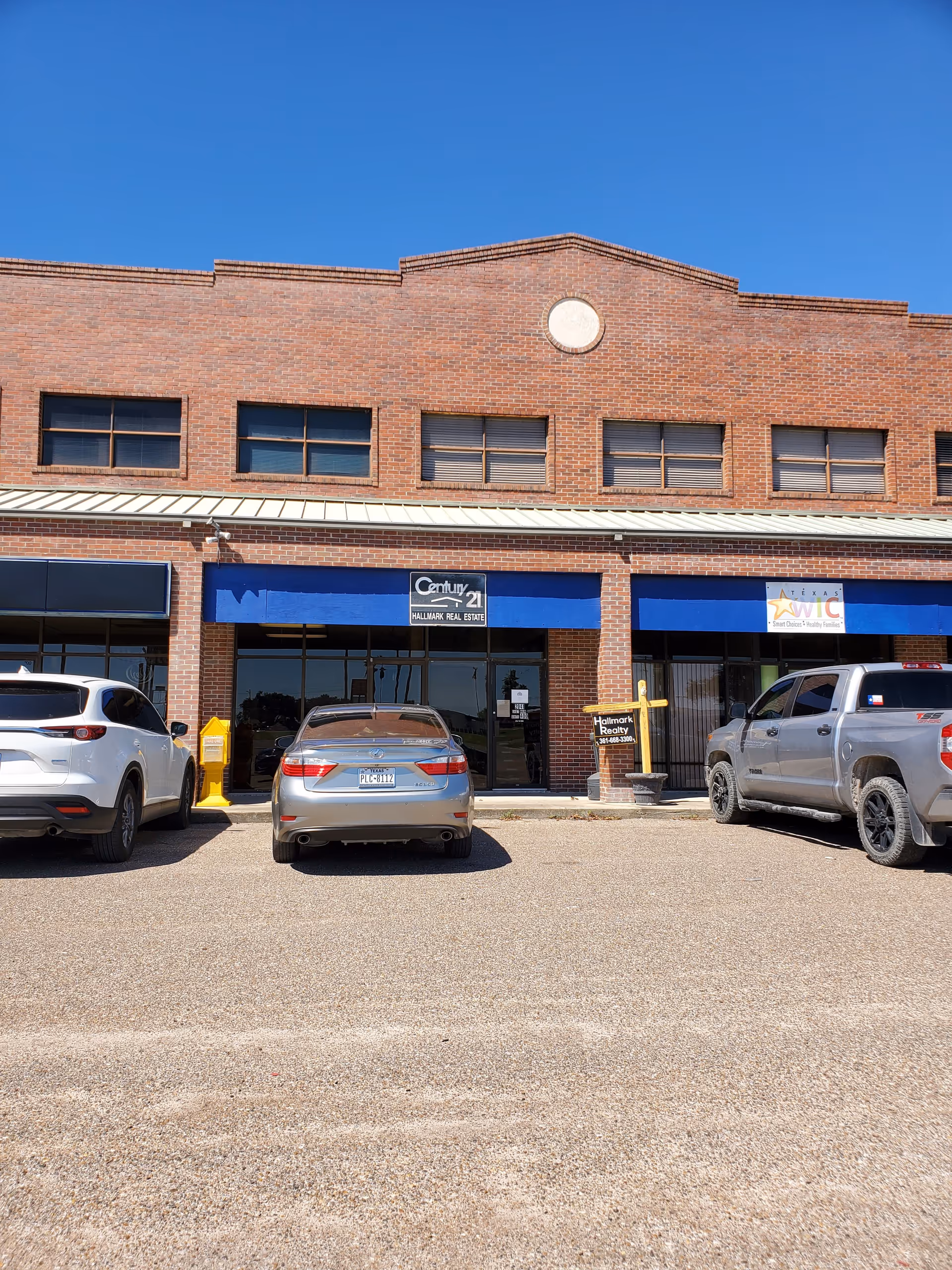 Brick storefront with blue awnings and multiple parked cars in front of a strip mall.