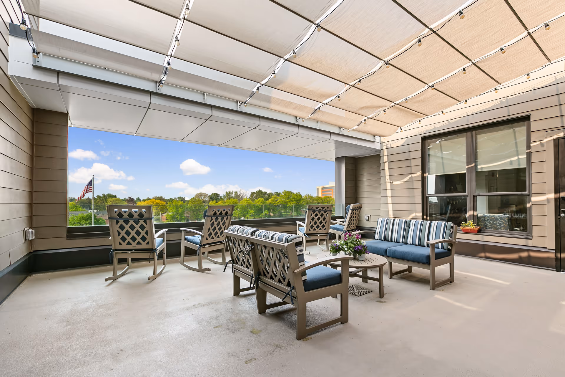 Outdoor terrace with cushioned seating, tables, string lights, and a leafy view.