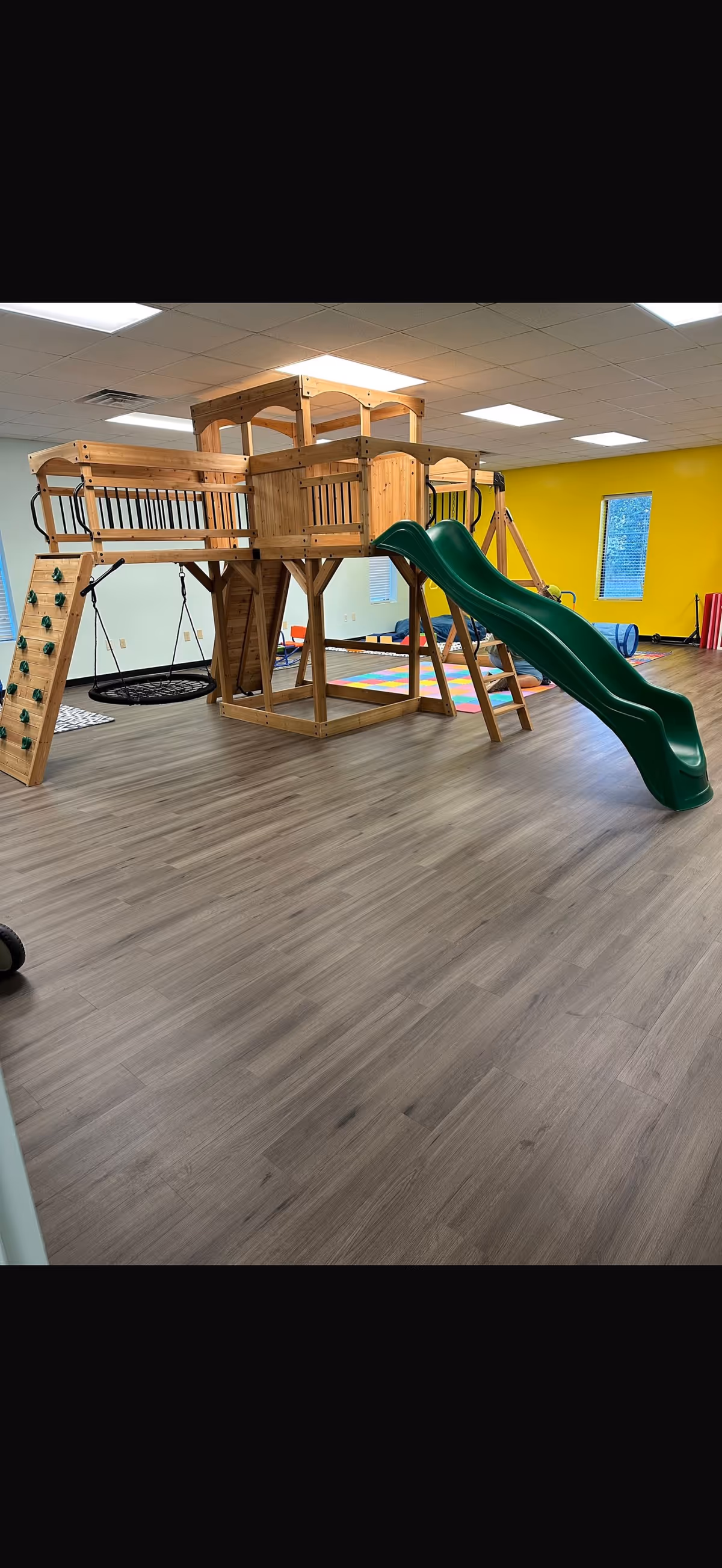 Indoor children's play structure with a wooden climbing frame and a green curved slide.