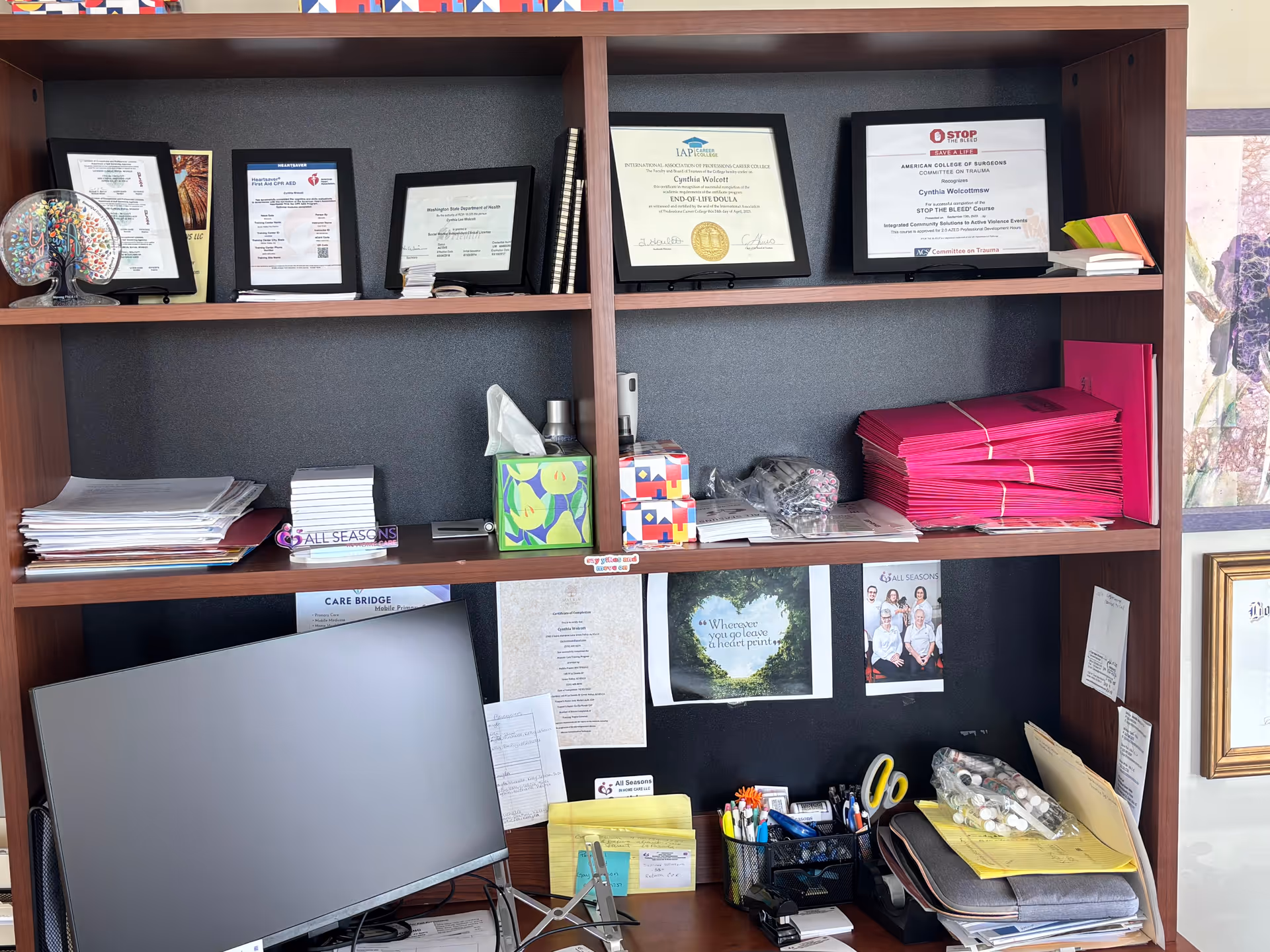An organized office workspace with certificates, a computer monitor, and supplies in a care facility.