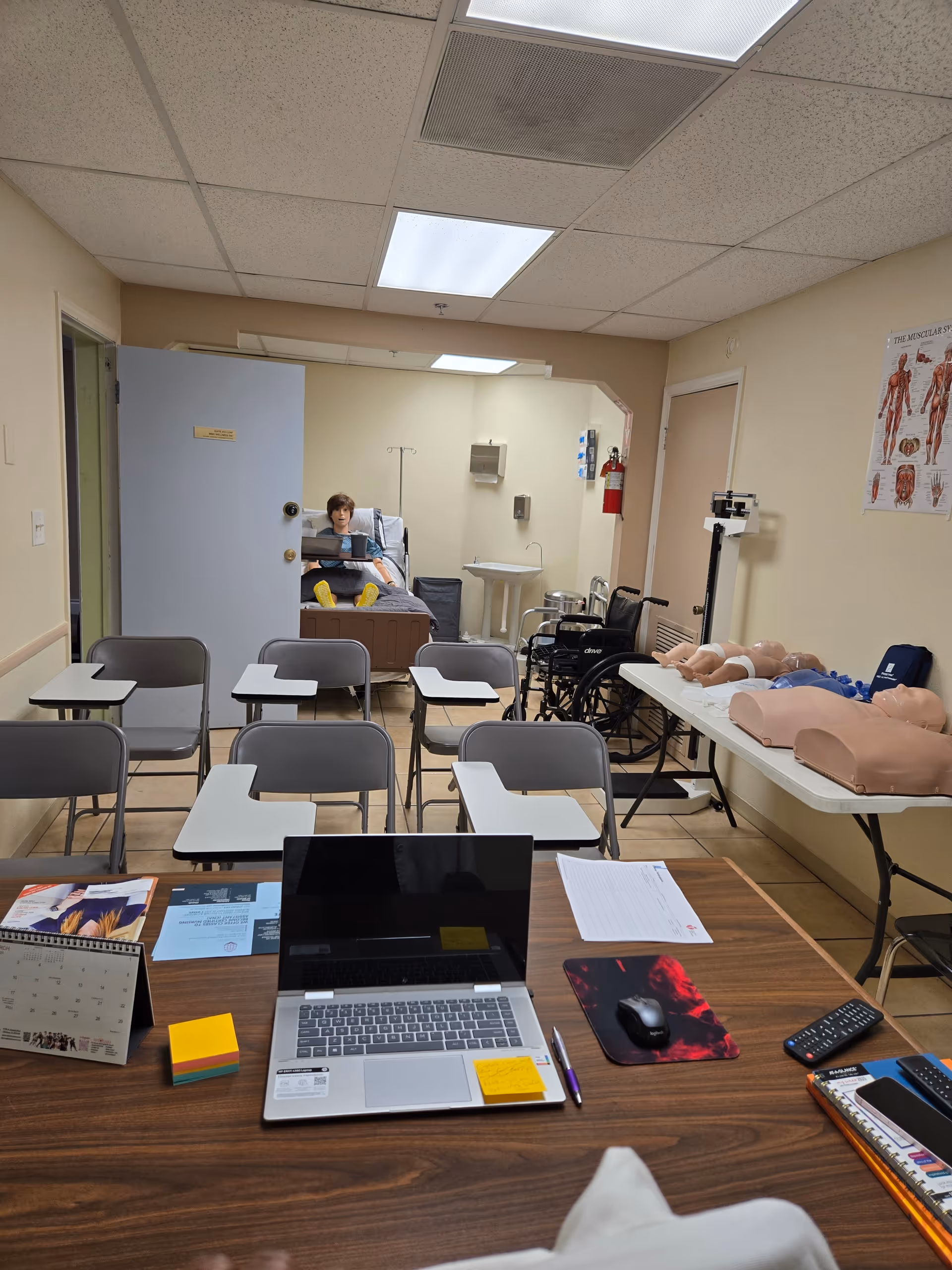 Classroom-style training room with medical mannequins, wheelchairs, and a laptop.