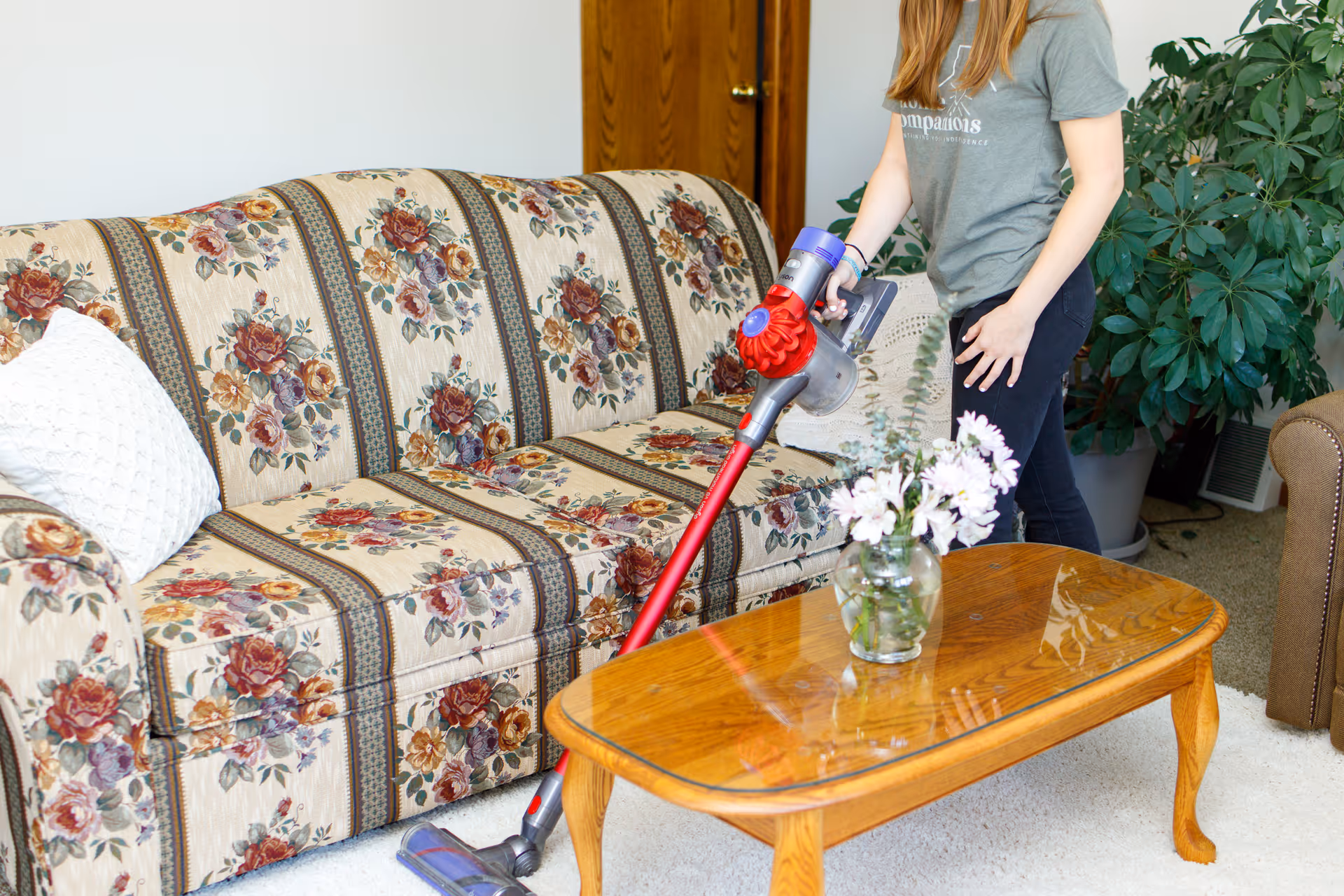 A person uses a cordless stick vacuum on a floral sofa in a living room.