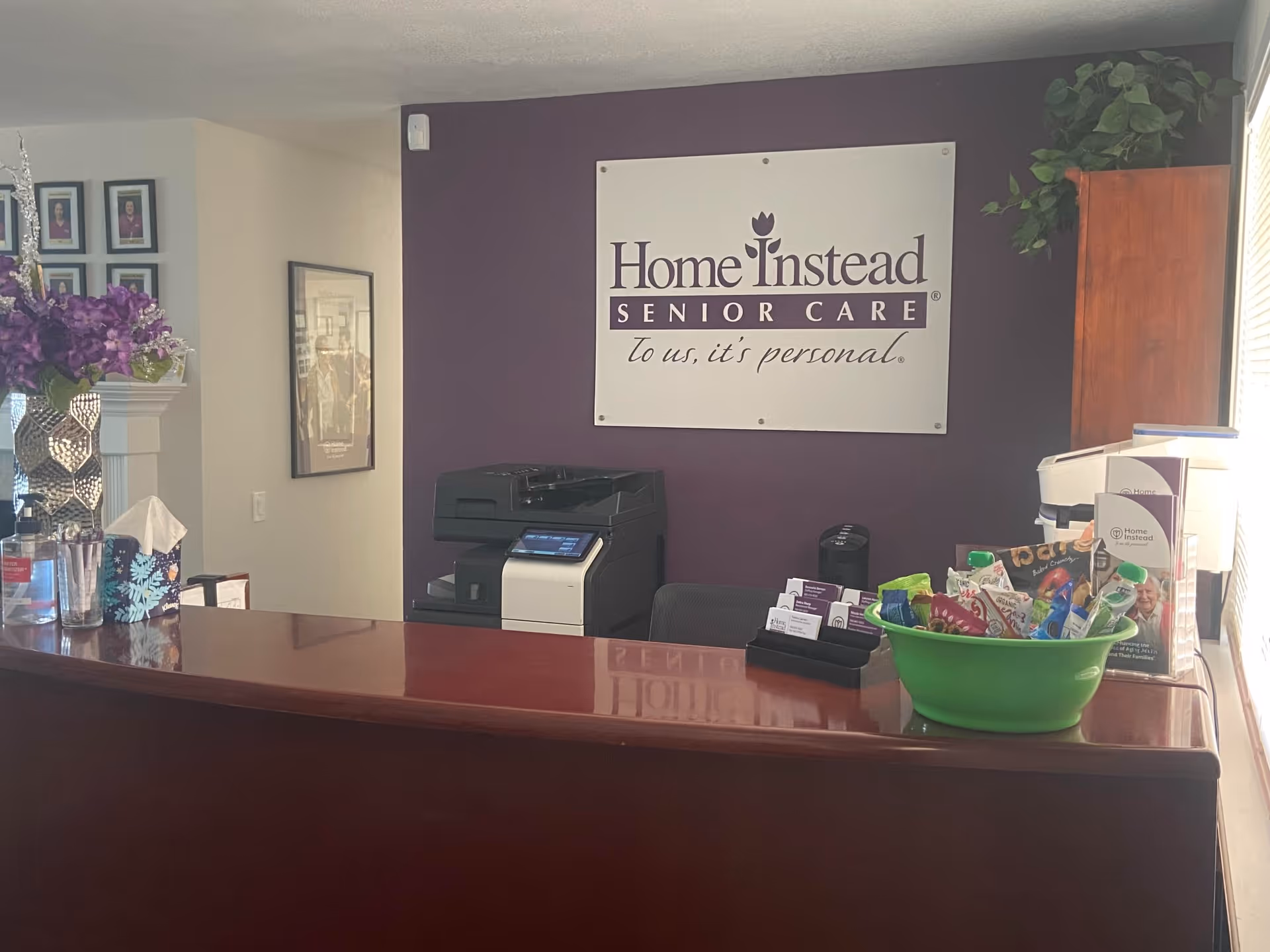 Reception desk with Home Instead sign, printer, and brochure bowl inside a purple-walled lobby.