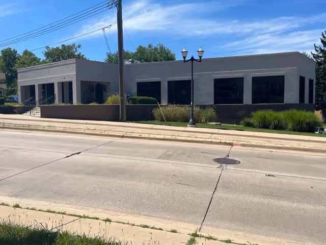 Gray single-story building with large windows along a street, landscaped with shrubs.