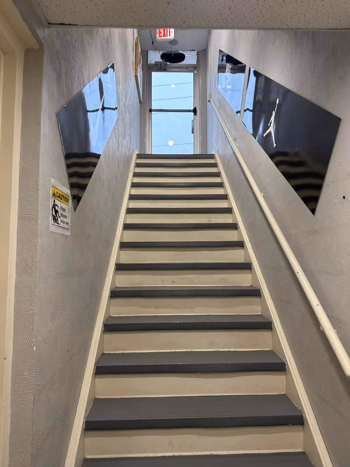 Narrow indoor stairwell with gray walls, blue-gray steps, handrail, and exit sign.
