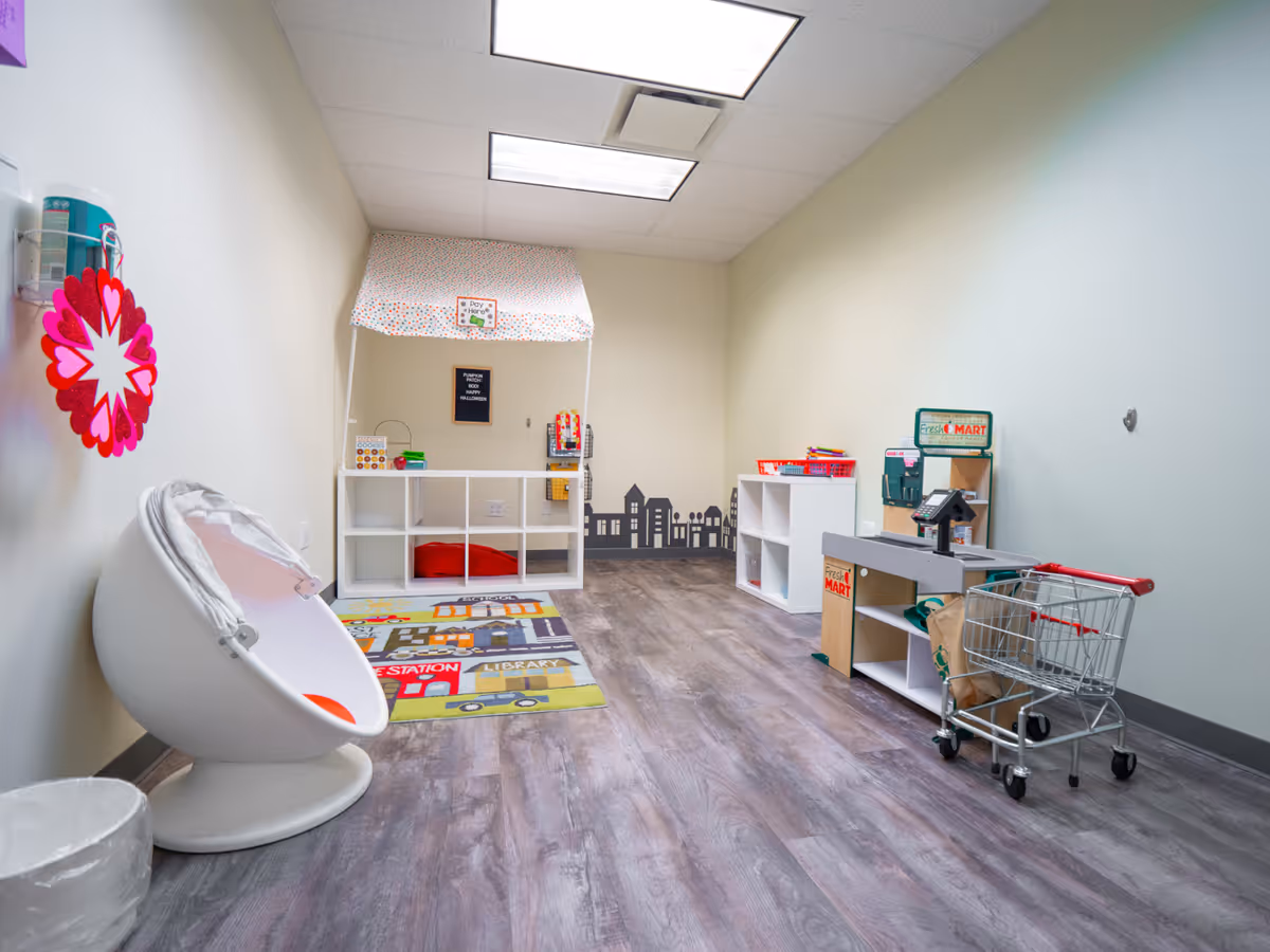 Child-friendly therapy room with shelves, a play market, shopping cart, and colorful road rug.