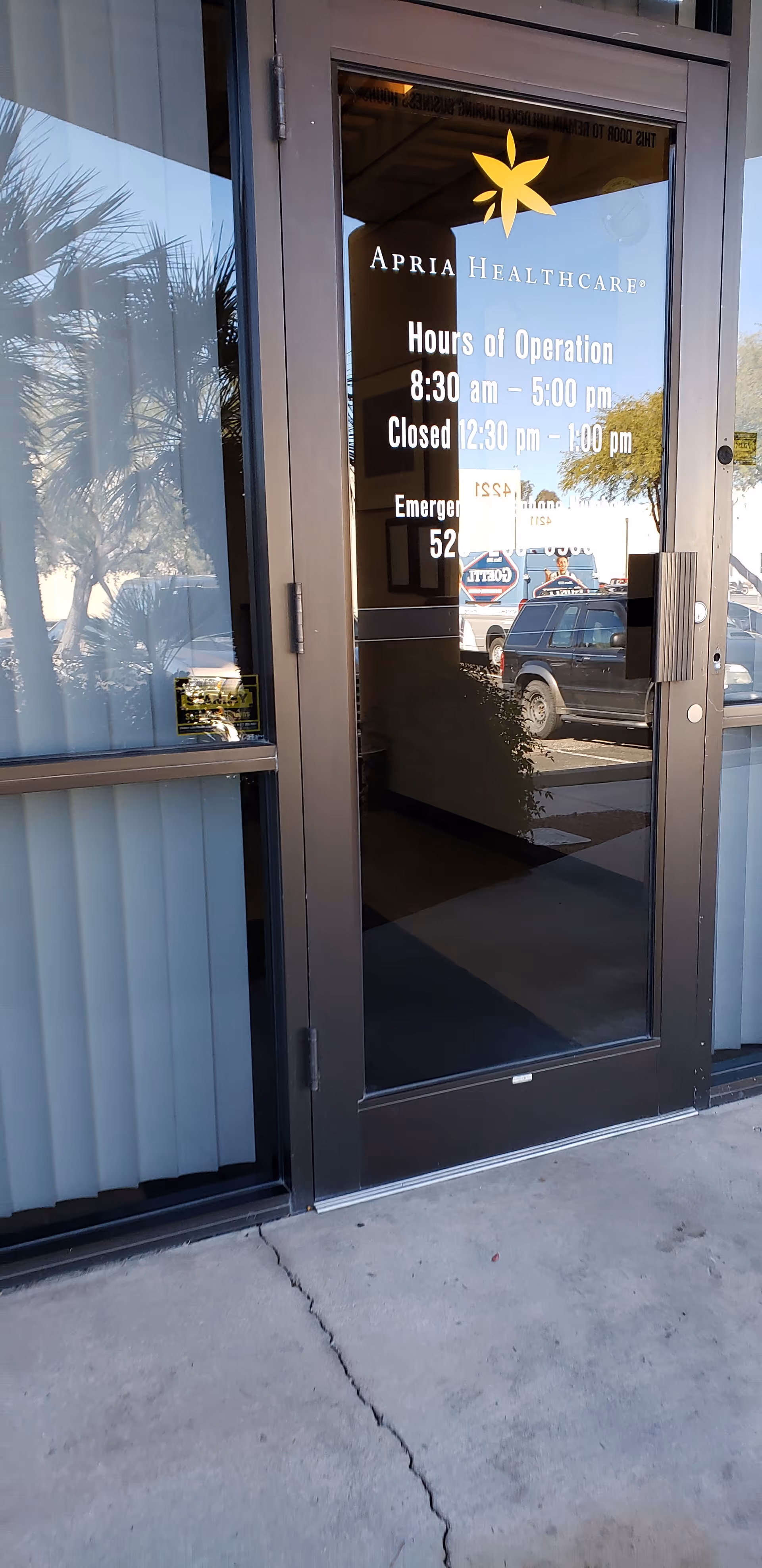 Glass entrance door displays Apria Healthcare hours with reflection of parked cars