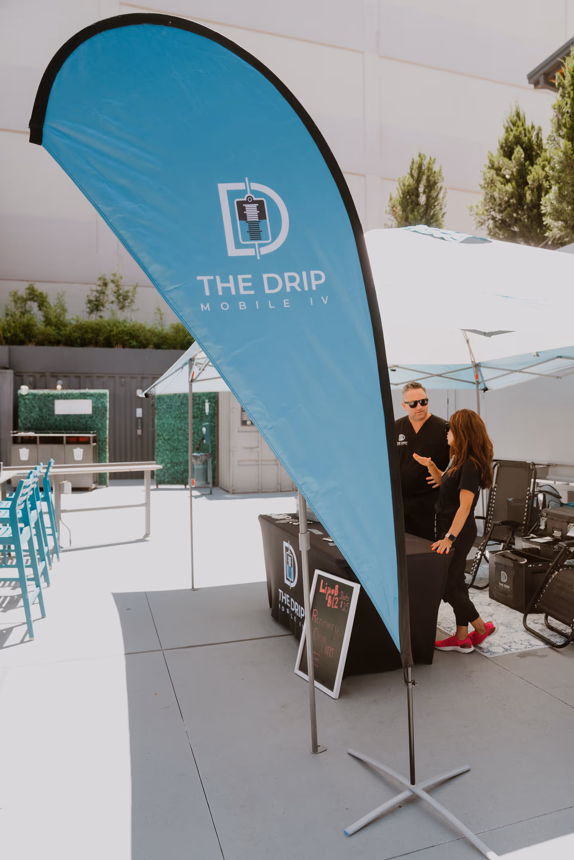 A blue The Drip Mobile IV flag stands beside a tented outdoor booth with two people.