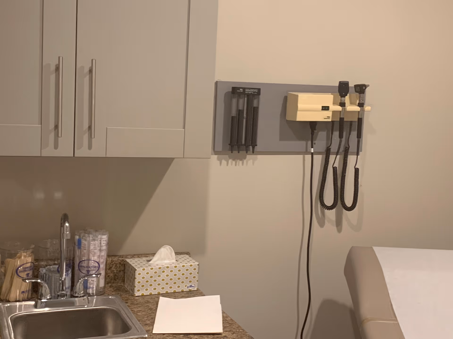 Clinic exam room with sink, tissue box, cabinets, and wall-mounted medical equipment.
