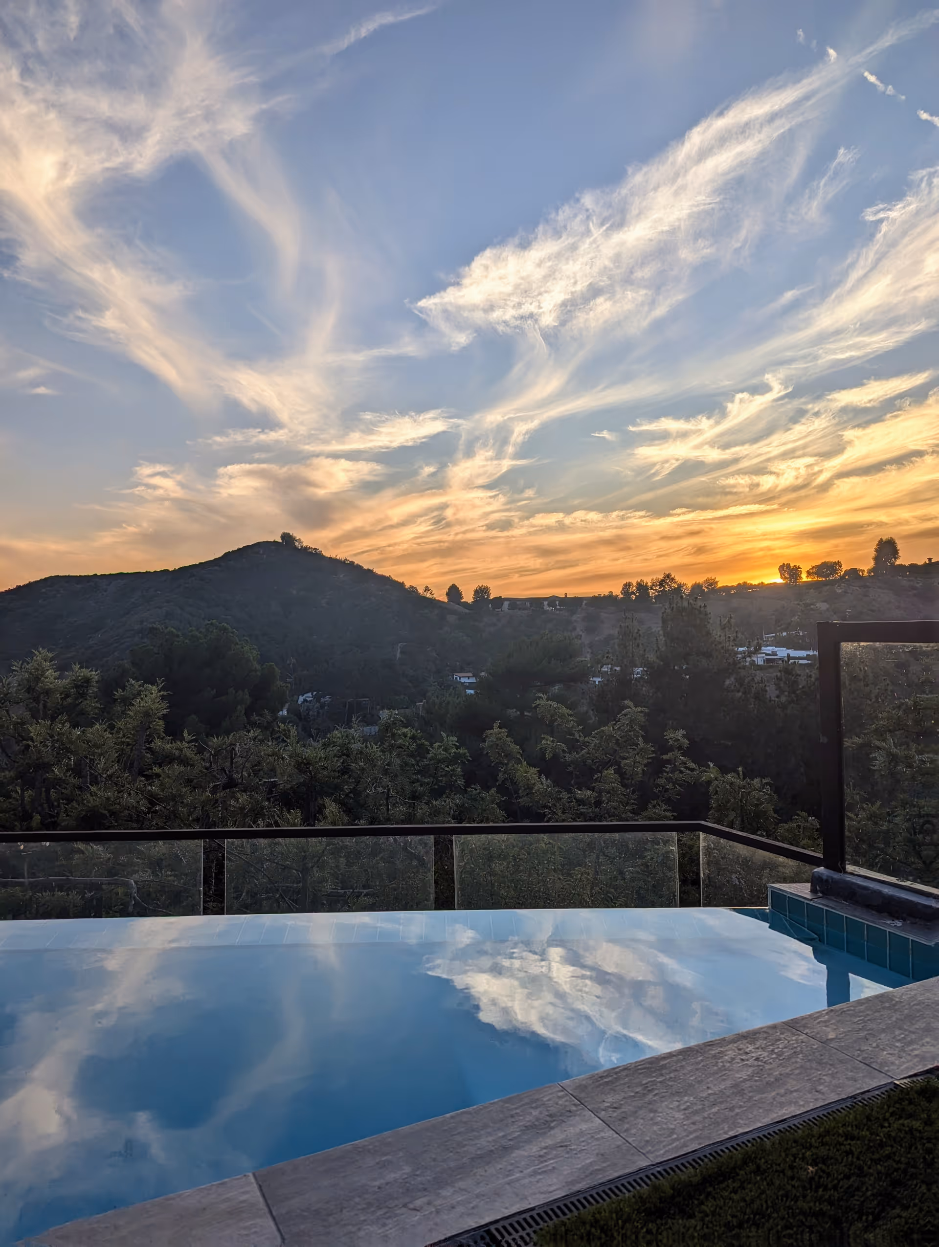 Sunset over distant hills seen from an outdoor pool deck with glass railing.