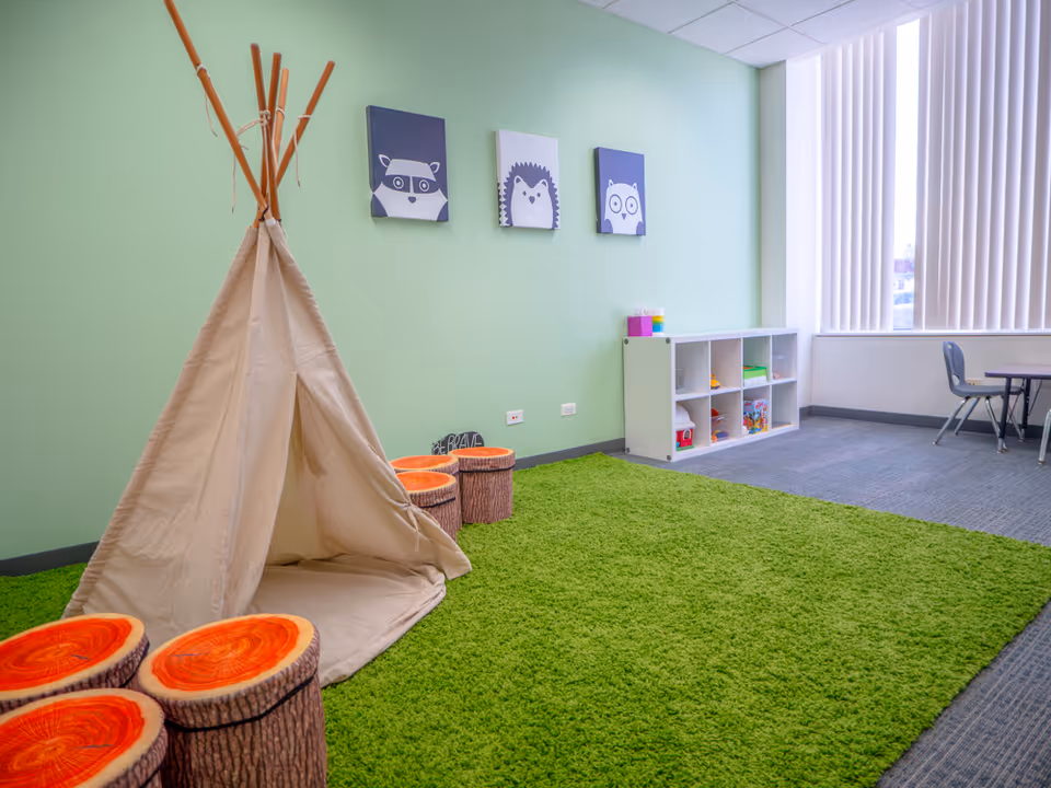 Bright indoor play area with a beige teepee, stump seating, green rug, and toy shelf.