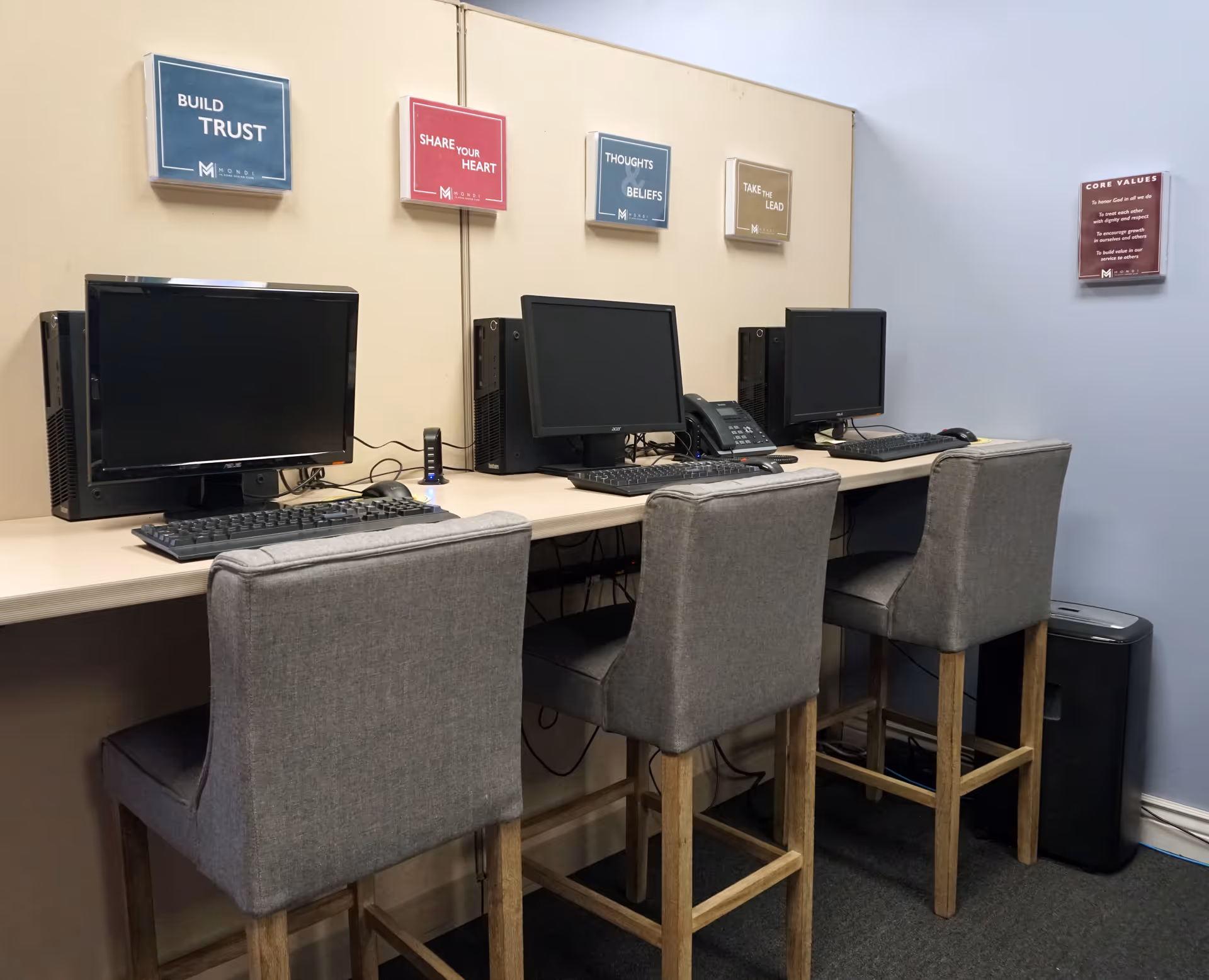 A computer workstation with three desktops and gray chairs in a senior living facility.