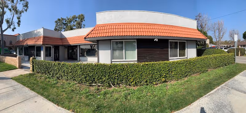 Exterior of a single-story building with red-tiled awning, hedges, and sidewalk.