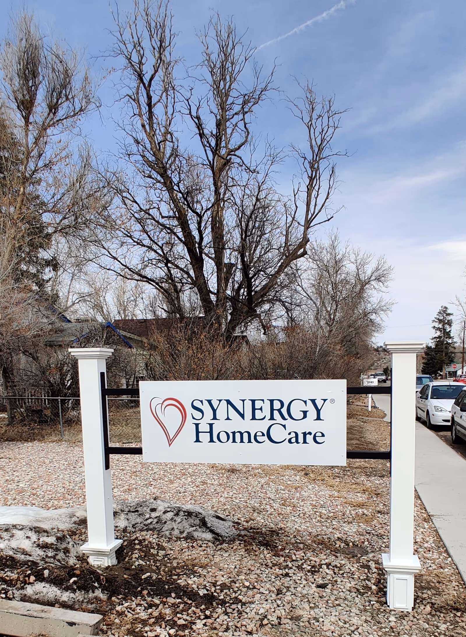 Synergy HomeCare sign on white posts amid gravel and bare trees.