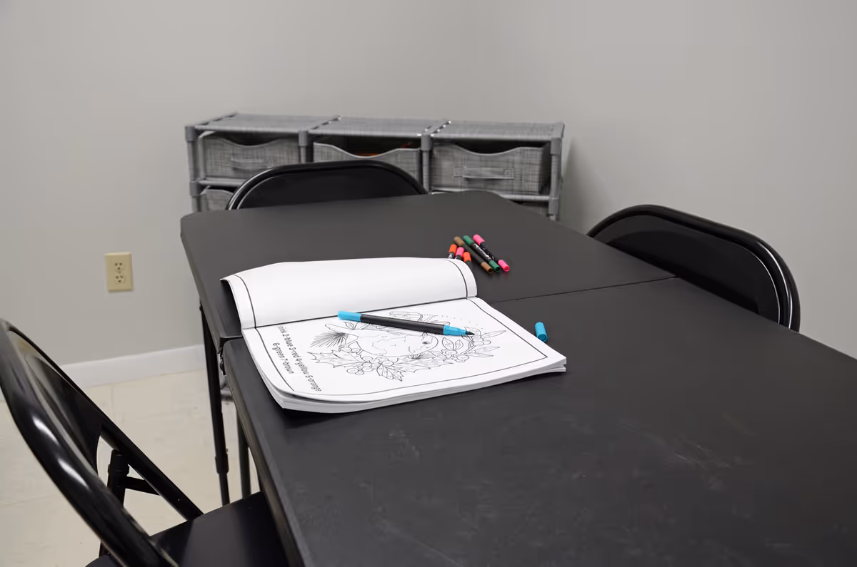 Dark craft table with coloring pages, markers, and storage bins in a plain room.