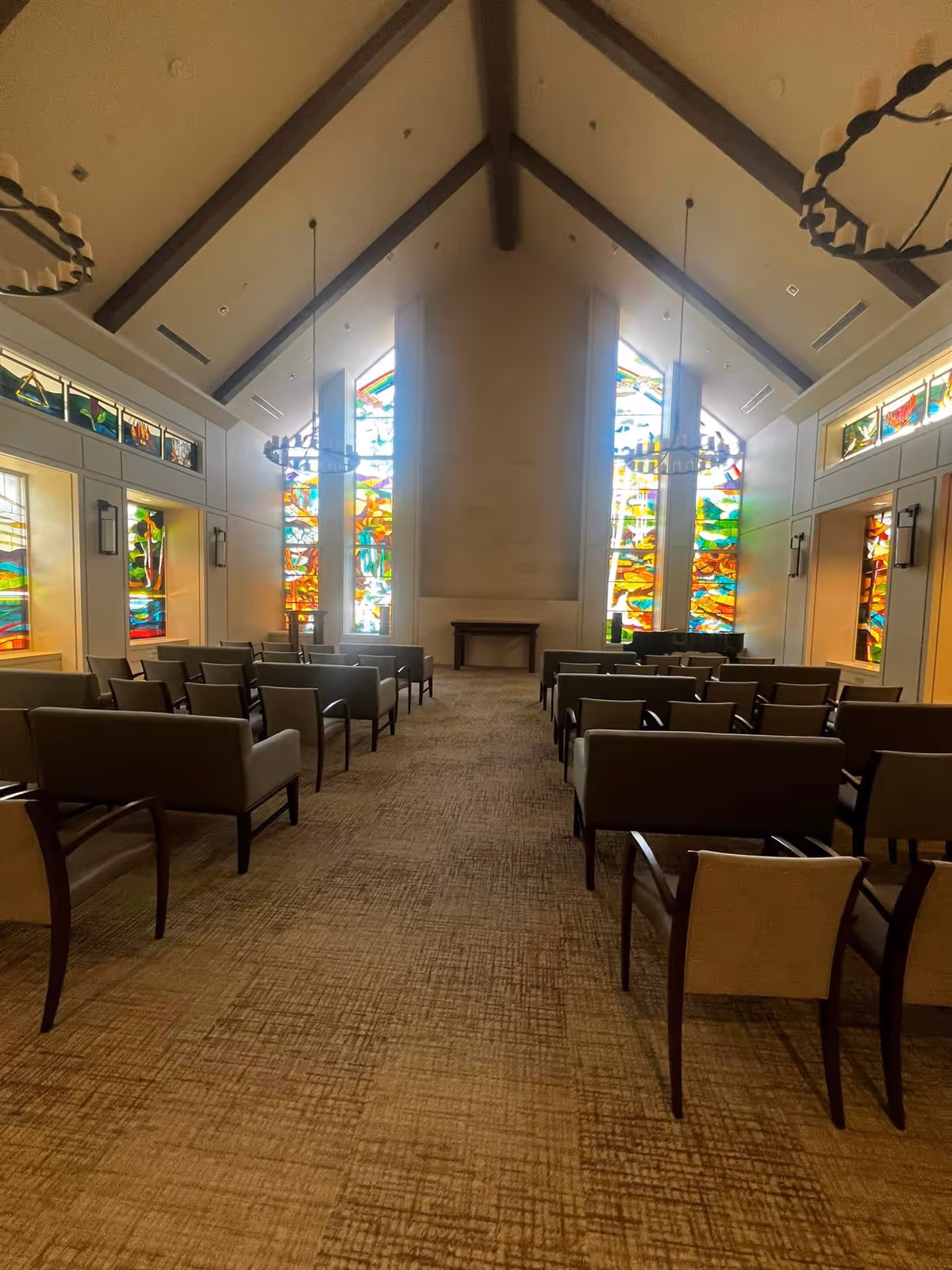 Interior chapel with rows of chairs, high ceiling, and colorful stained-glass windows at front.